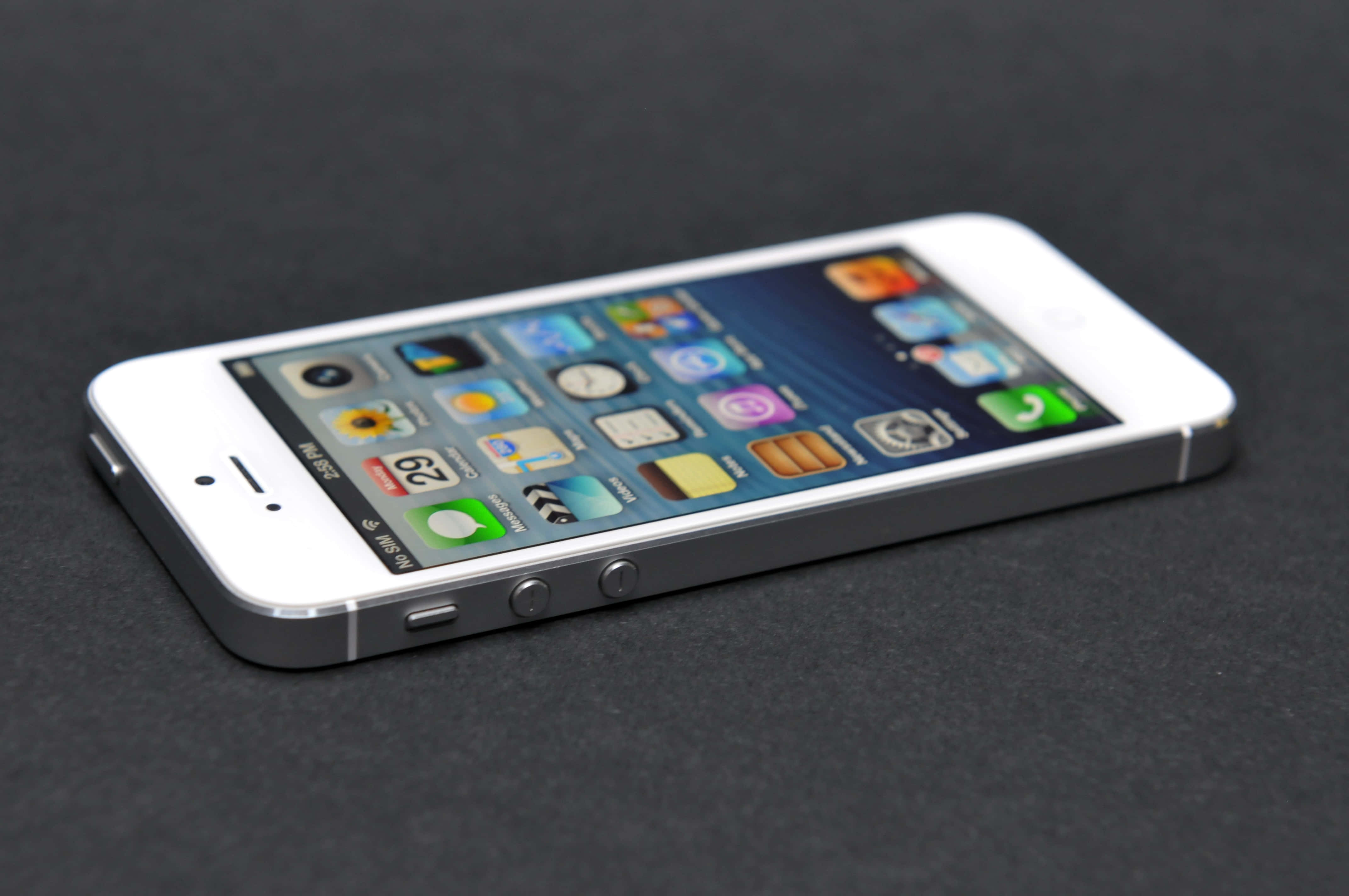 Apple Phone 桌布 壁紙 — 一部白色 iPhone 5s 傾斜放置在深色紋理表面上，其色彩繽紛的應用程式圖標在柔和燈光下發光。