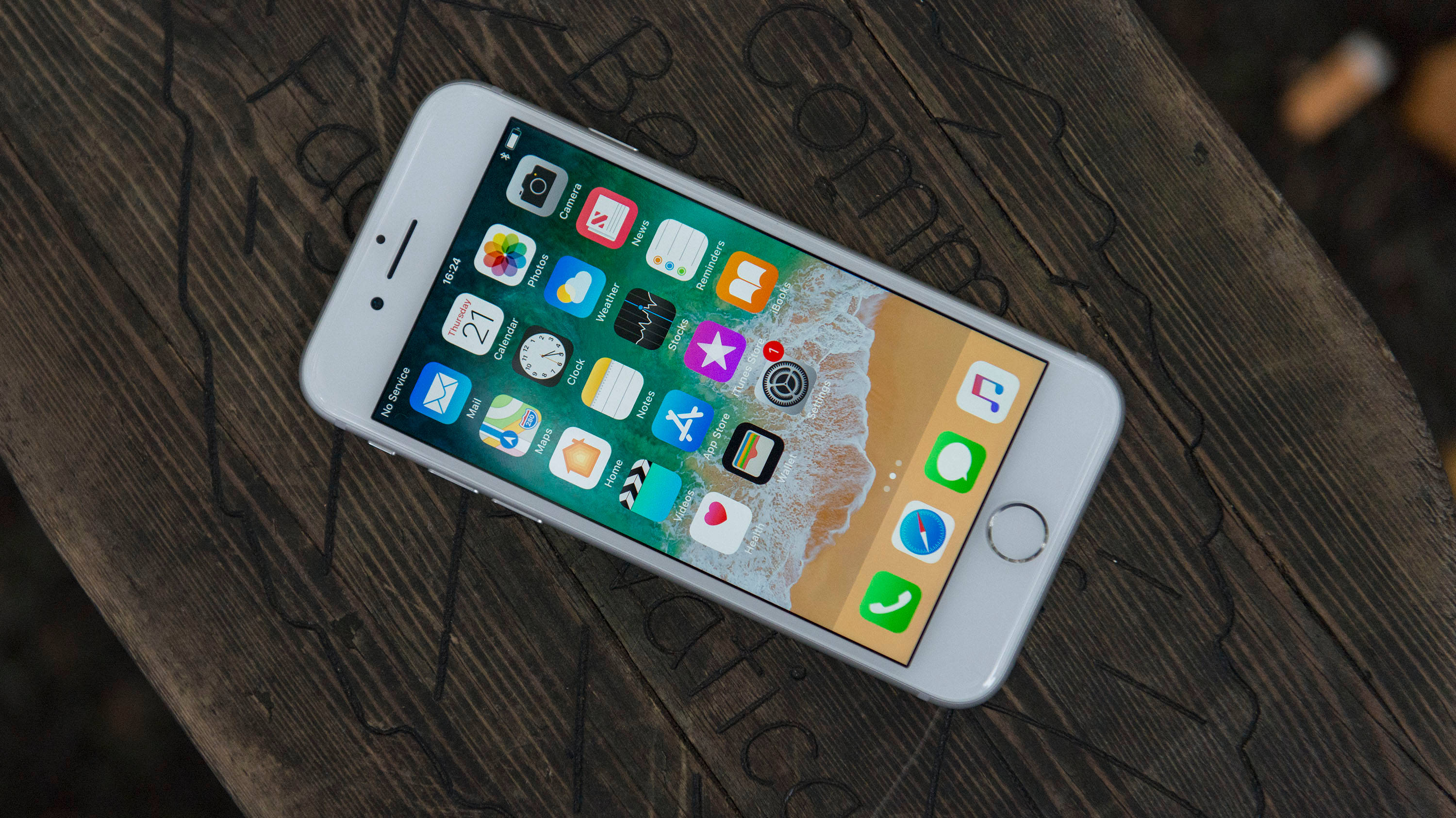 iPhone 8 桌布 壁紙 — 一部白色 iPhone 6s 放在深色木質表面上，顯示其色彩繽紛的應用程式圖示和海灘桌布。