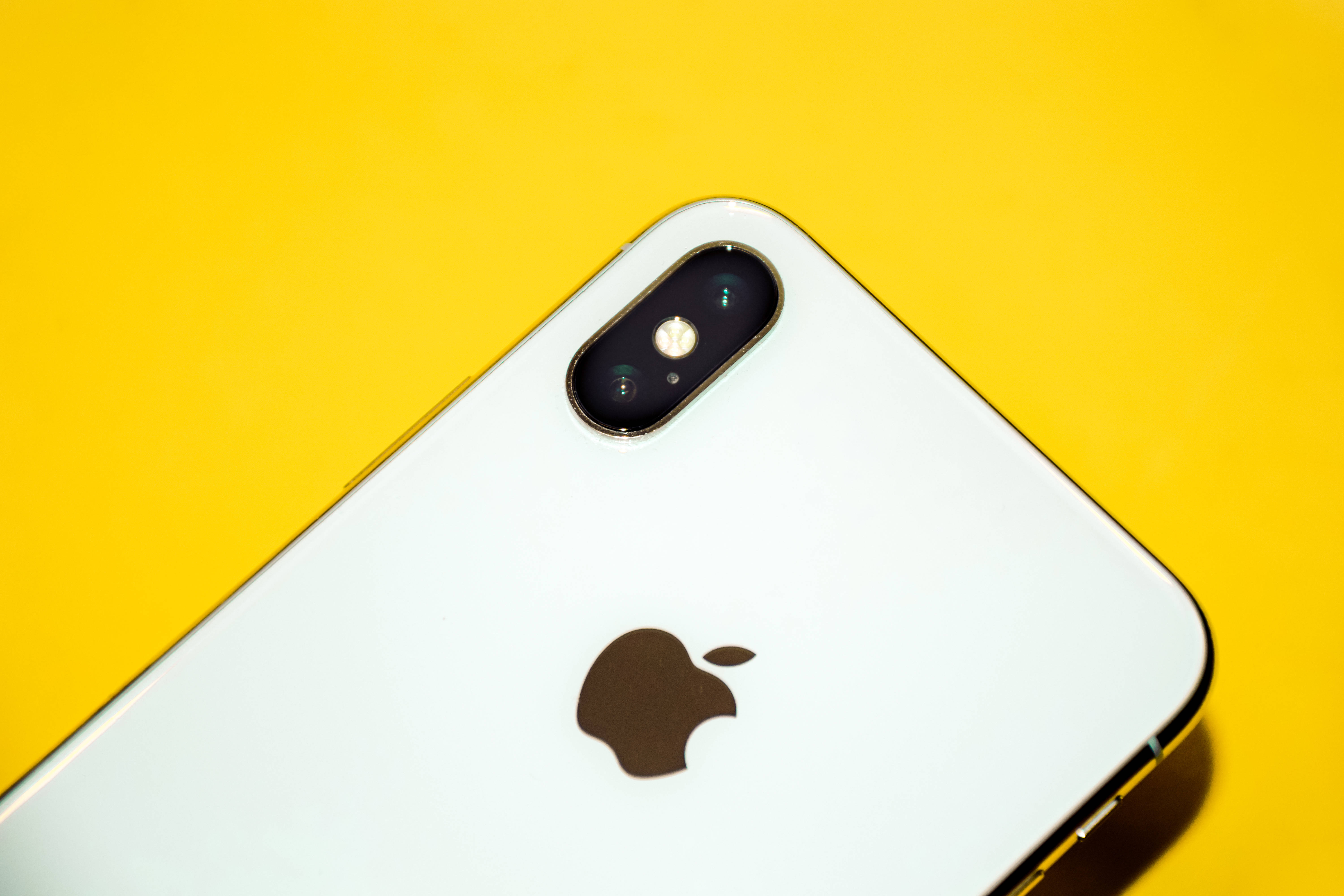 iPhone Xs Max 桌布 壁紙 — 一部白色iPhone，配備雙鏡頭和蘋果標誌，傾斜放置在鮮黃色背景上。