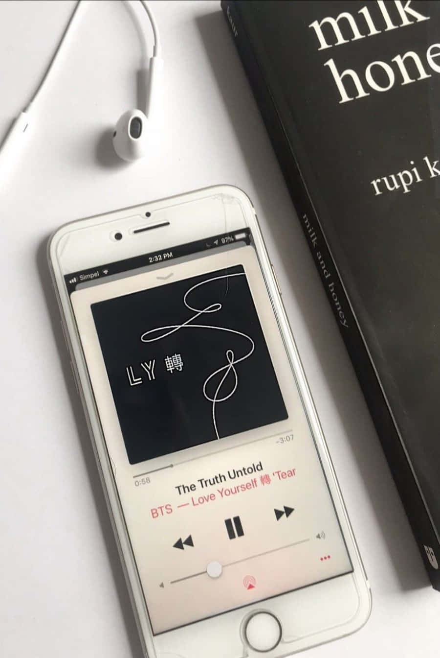 手機 桌布 壁紙 — 一部白色iPhone在播放BTS音樂，旁邊放著一本黑色《milk and honey》書本和白色耳機，置於簡約白色表面上。