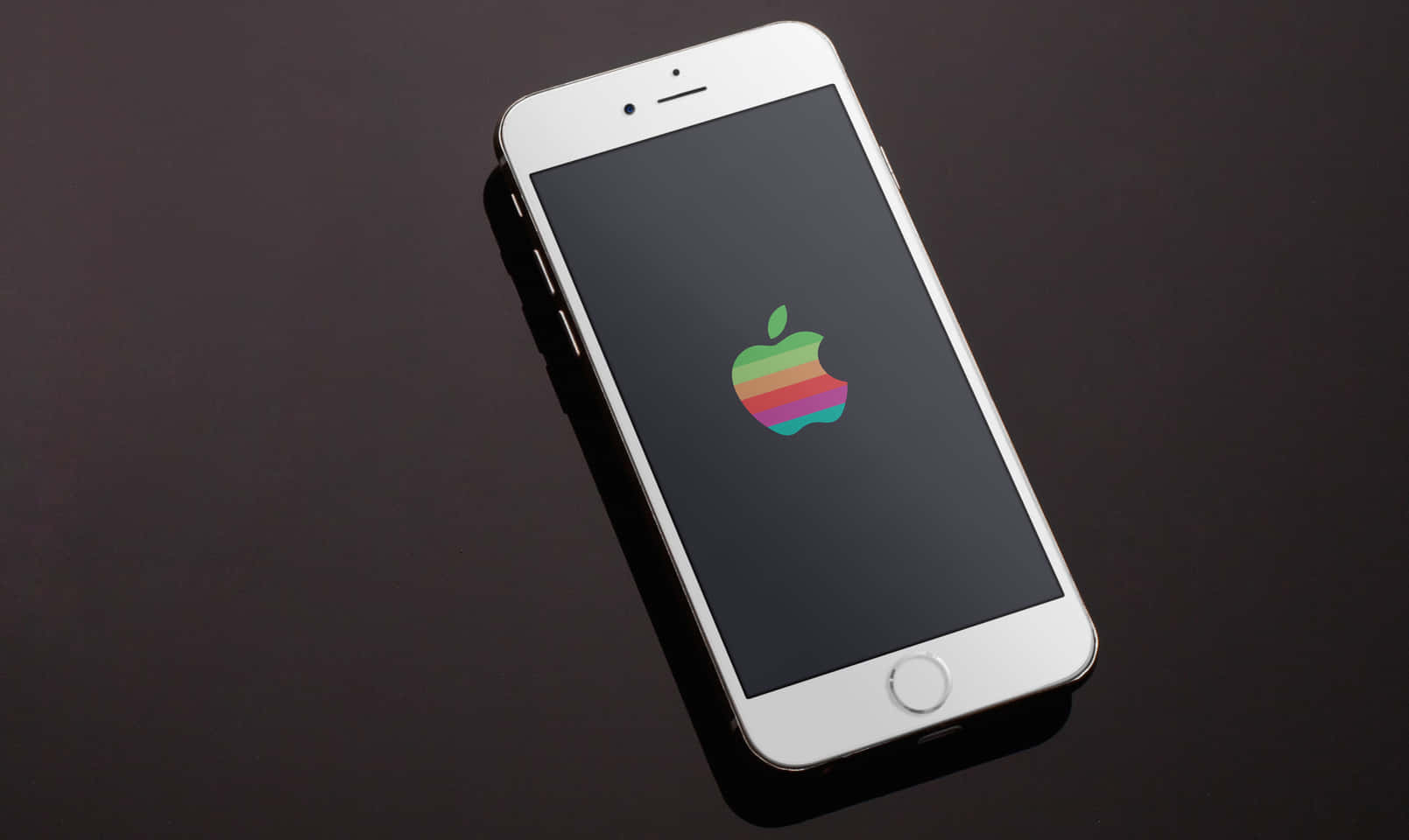 桌布 壁紙 — 一部白色iPhone在深色反光表面上顯示復古彩虹Apple標誌。