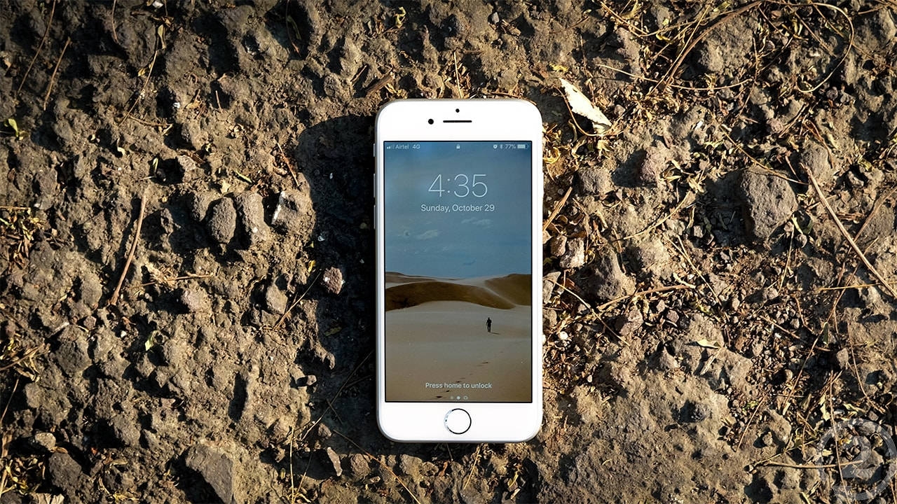 iPhone 8 桌布 壁紙 — 一部白色智慧型手機放在粗糙的泥土地上，顯示沙漠桌布，其中一個孤獨的人影在沙丘之間行走。