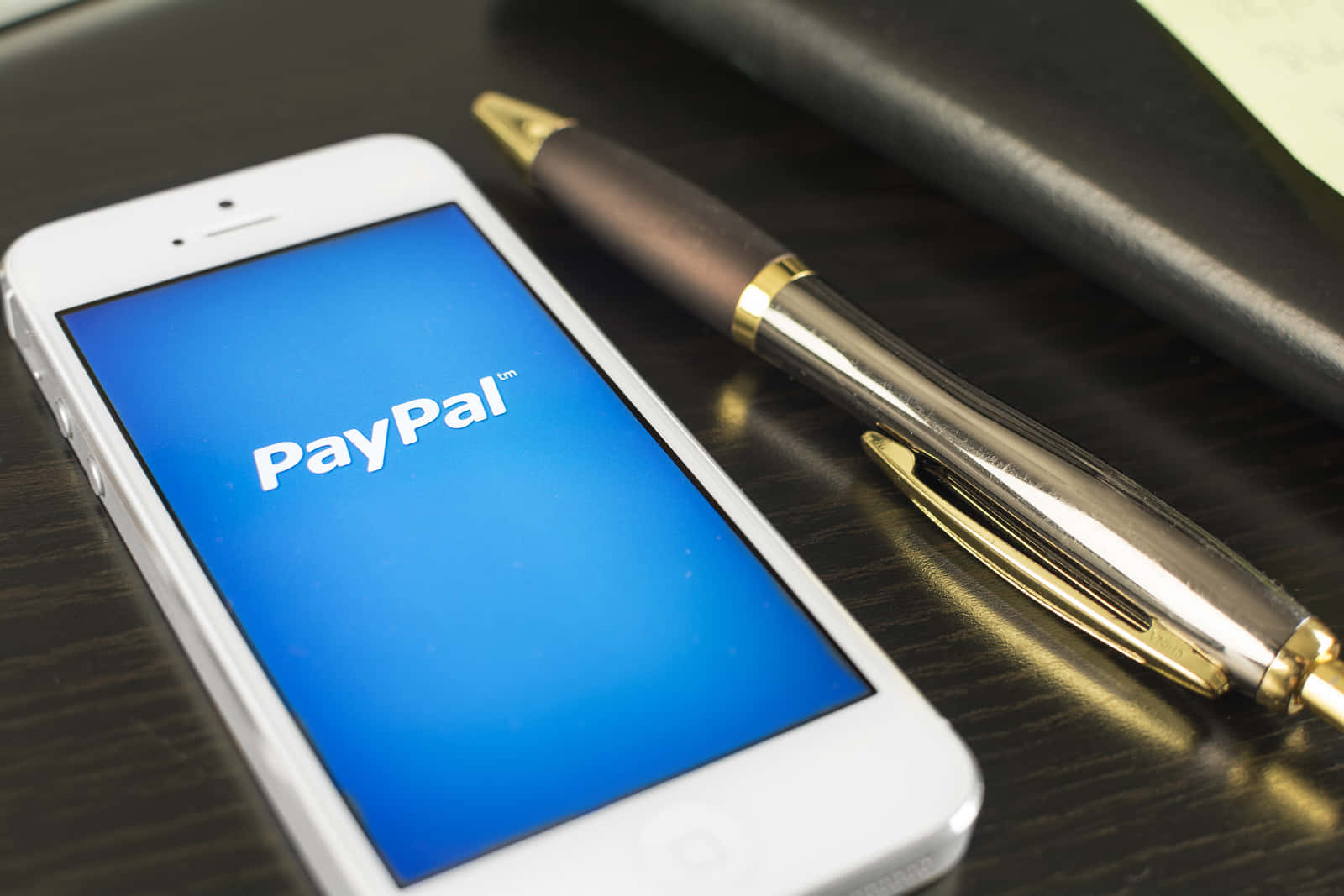 PayPal 桌布 壁紙 — 一部白色智慧型手機顯示PayPal標誌，旁邊放著一支金屬筆在深色木桌上。