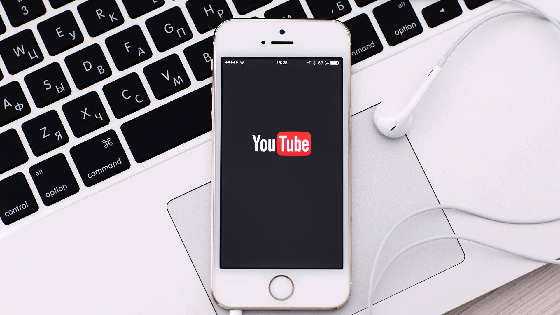 YouTube Logo 桌布 壁紙 — 一部白色智慧型手機顯示YouTube標誌，放在筆記型電腦鍵盤上，旁邊有白色耳機。