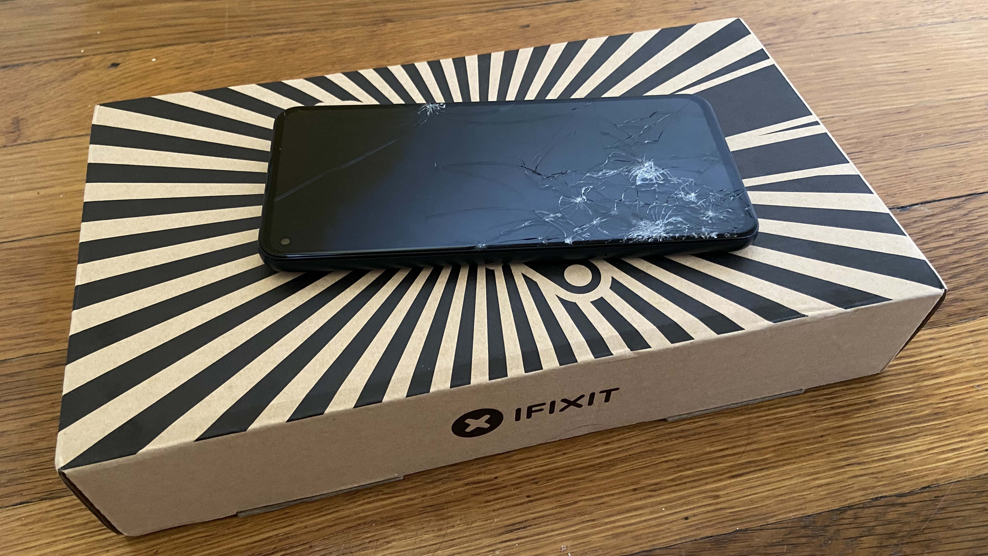 裂屏 桌布 壁紙 — 一部破裂的黑色智慧型手機放在木桌上一個黑色和米色條紋的 iFixit 盒子上。