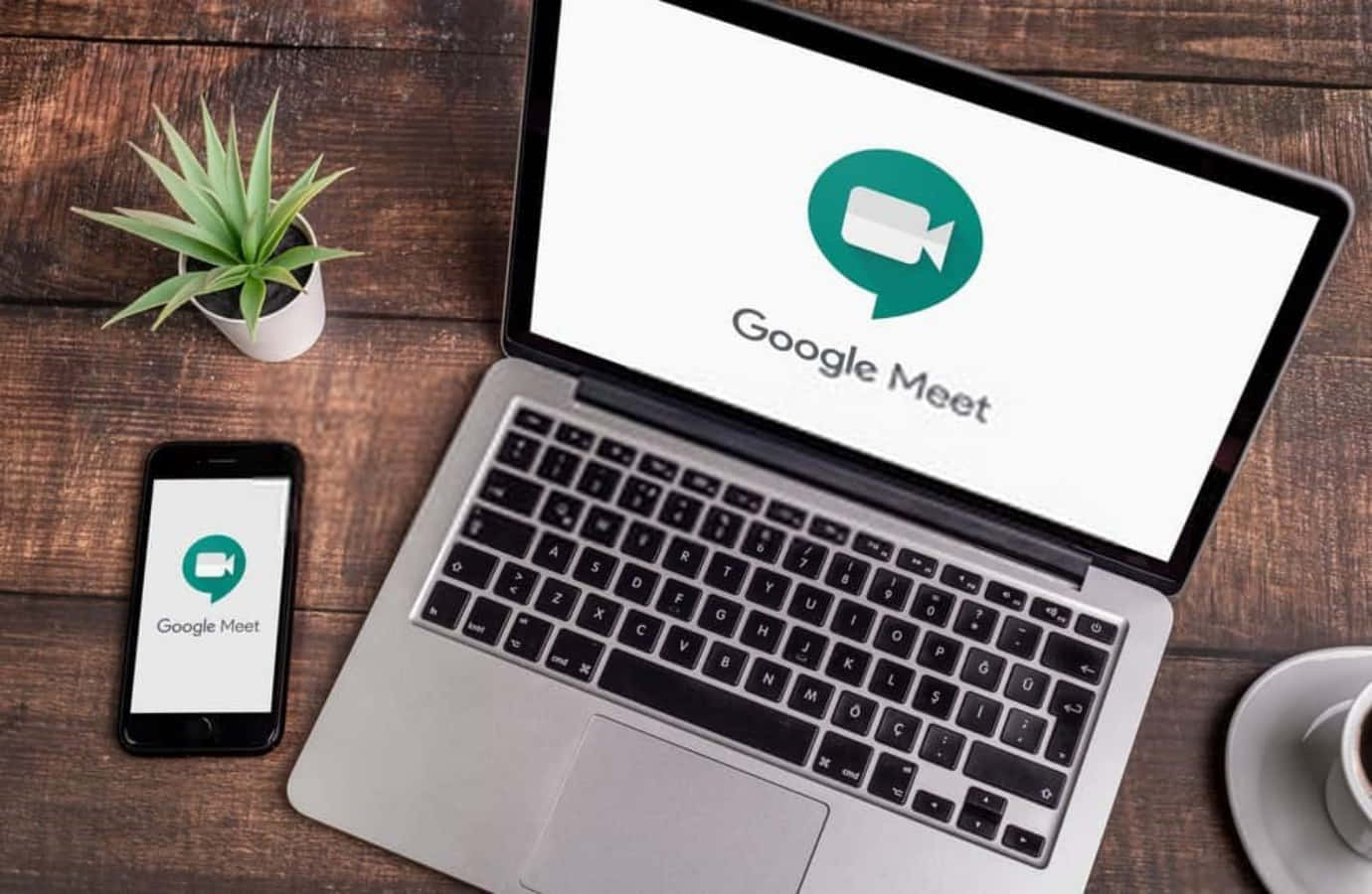 筆記型電腦和手機在木桌上顯示 Google Meet 應用程式，伴有植物和咖啡。 — Google Meet 壁紙