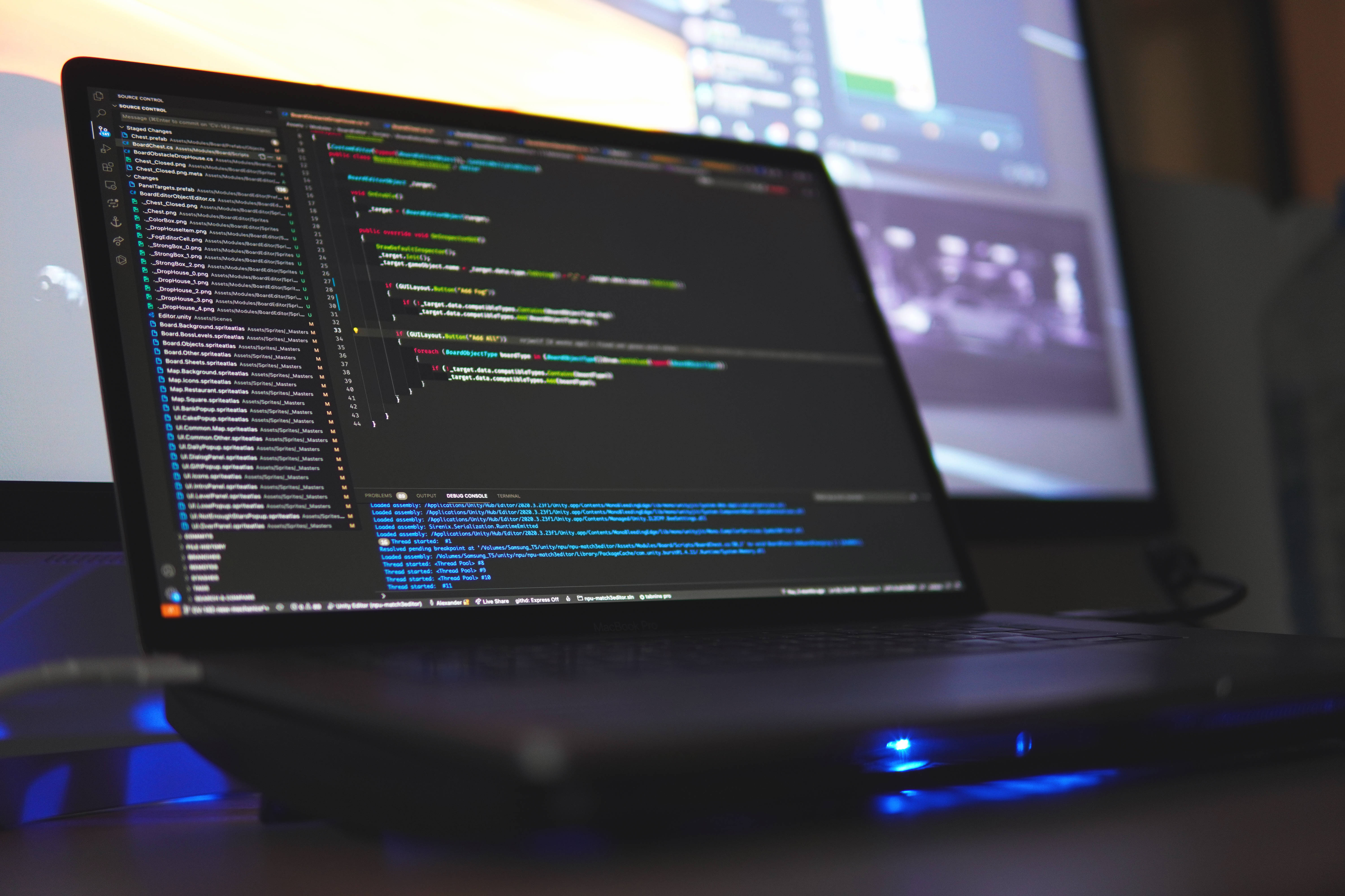 Coder 桌布 壁紙 — 一部筆記型電腦在暗色編輯器中顯示彩色程式碼，放在桌上，帶有發光的藍色燈光和背景中的第二個顯示器。