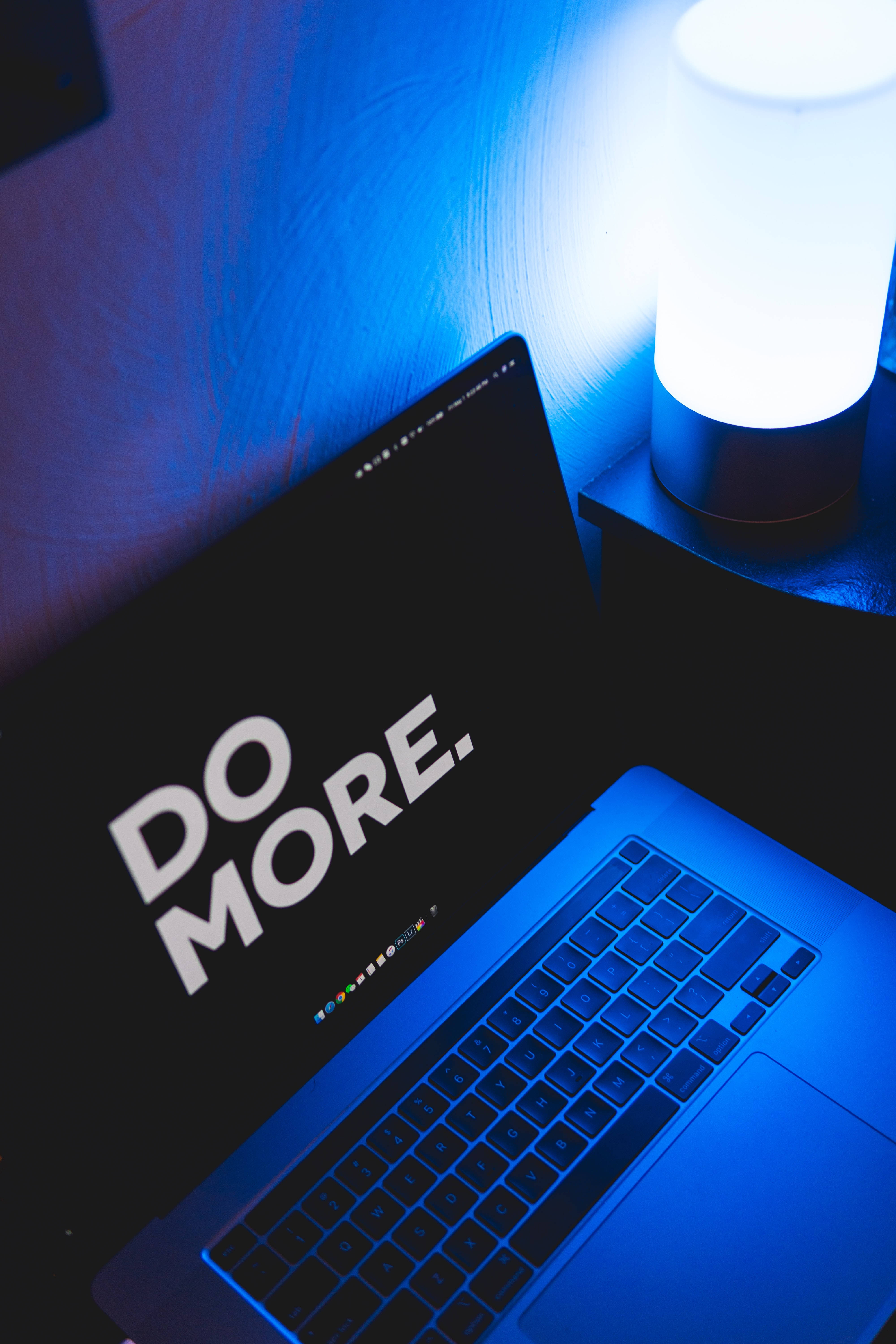 激勵 桌布 壁紙 — 一部筆記型電腦顯示 'DO MORE.' 以粗體白色文字，沐浴在旁邊發光燈泡的冷藍光中。