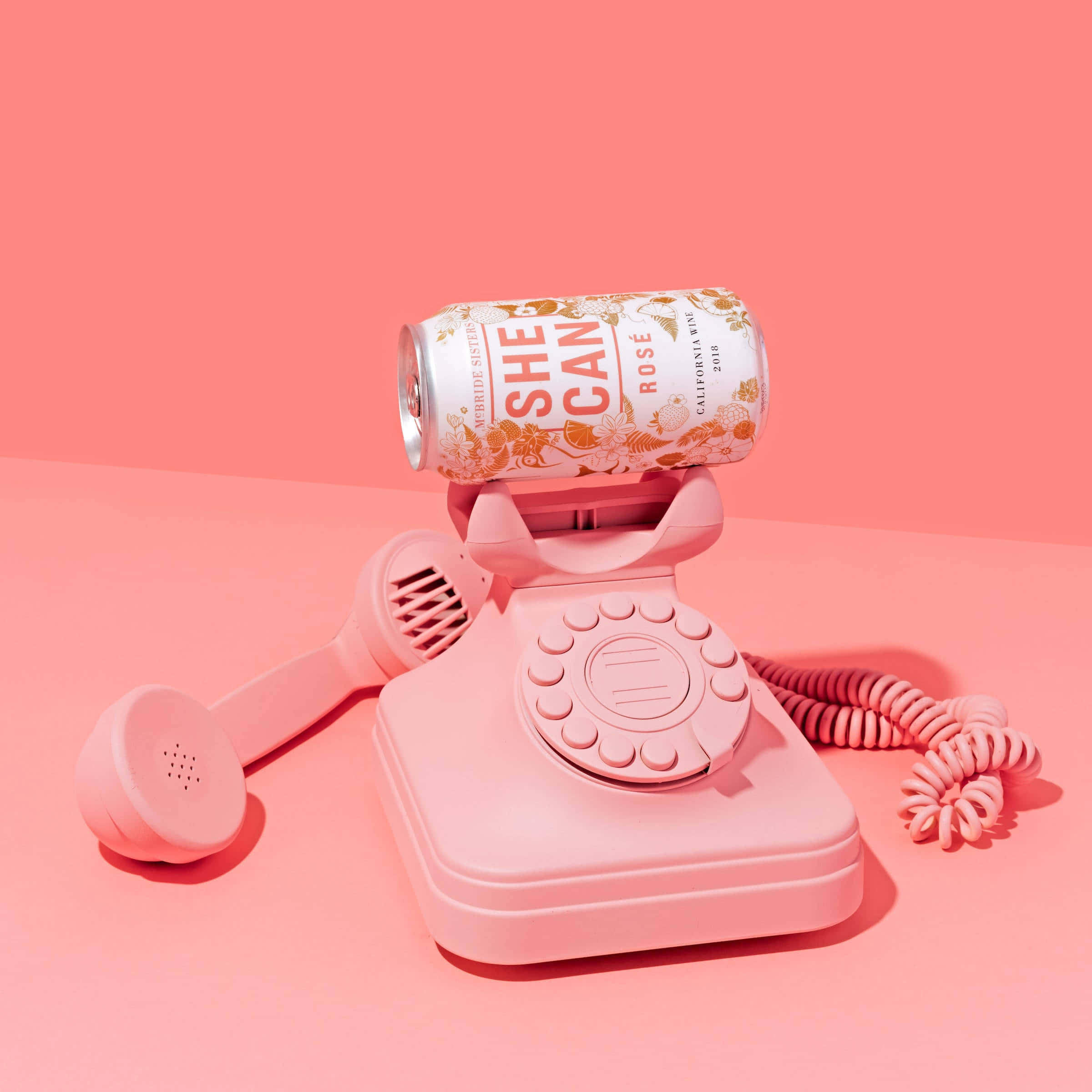 Telephone 桌布 壁紙 — 一部粉紅色轉盤電話機拿著一個相配的粉紅色葡萄酒罐，標籤為「She Can Rosé」，背景為純粉紅色。