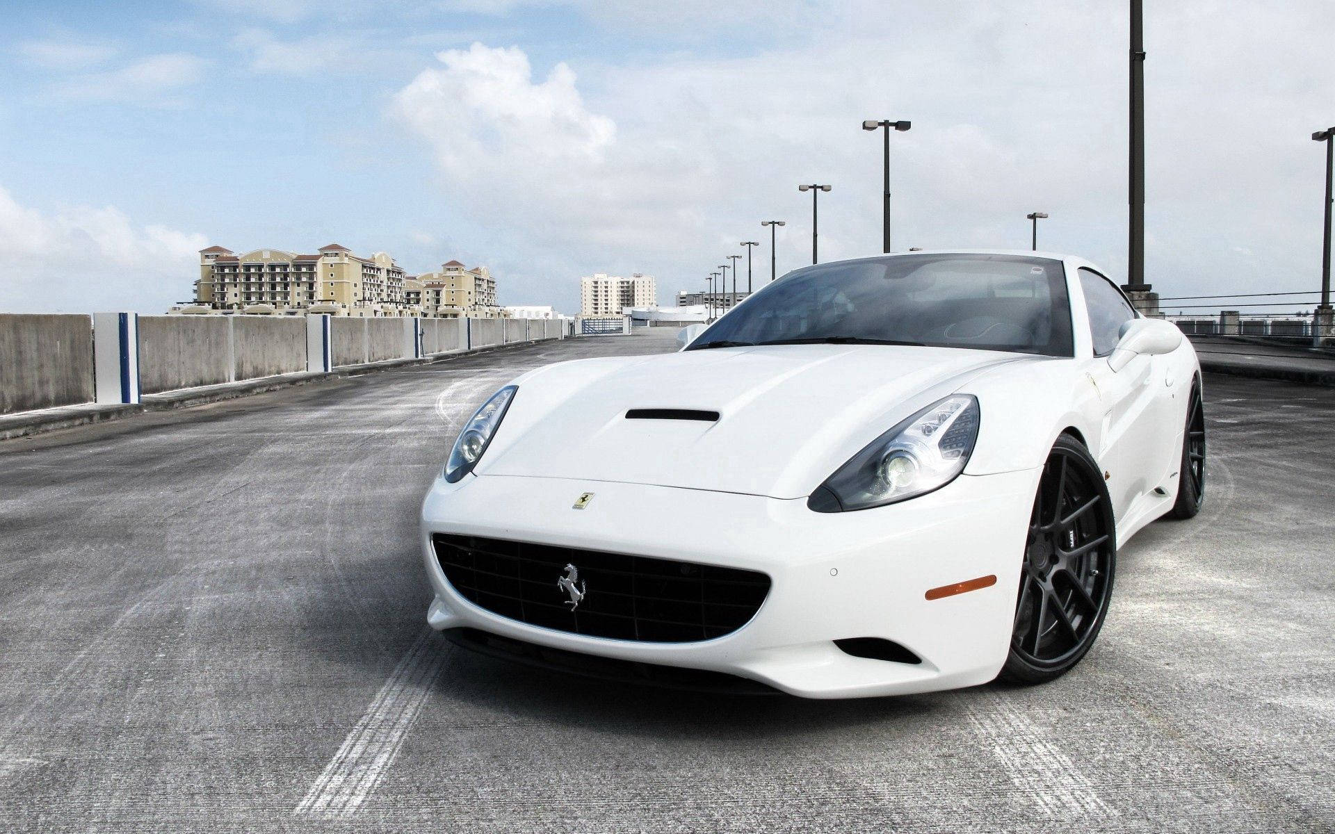 white ferrari 桌布 壁紙 — 一部純白色法拉利 California 跑車停放在高架混凝土道路上，遠方建築物在局部多雲的天空下。