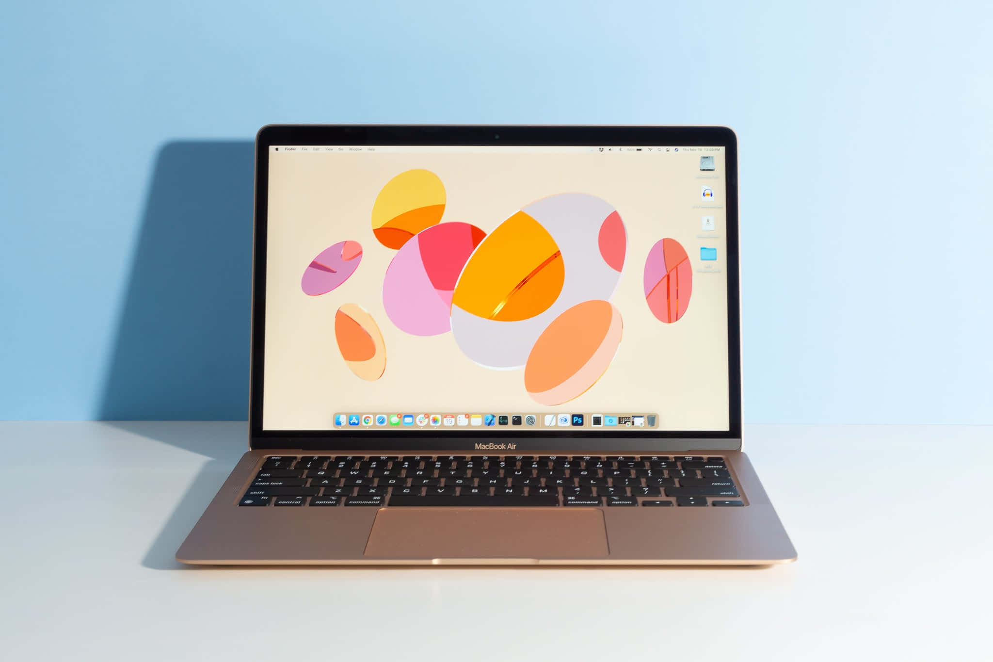 MacBook 動態圖片 桌布 壁紙 — 一部纖薄的金色 MacBook Air 在白色表面上打開，對著柔和的藍色牆壁，顯示充滿活力的抽象桌布，上面有重疊的彩色圓圈。