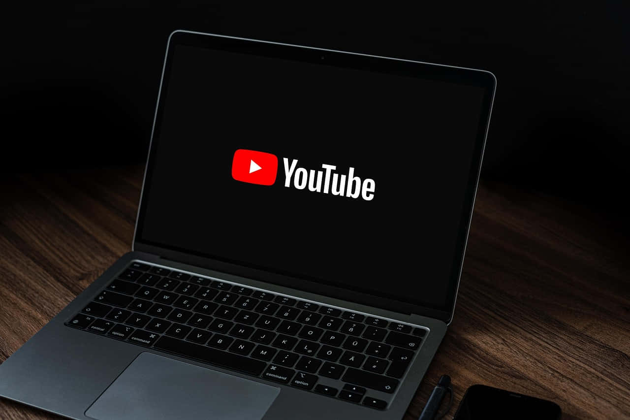 YouTube 桌布 壁紙 — 一部纖薄筆記型電腦在昏暗房間的木桌上，顯示YouTube標誌於黑色螢幕上。