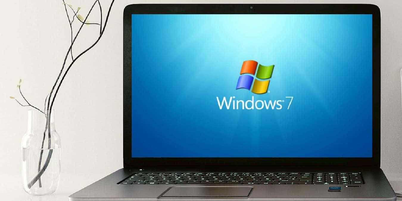 Windows 7 桌布 壁紙 — 一部纖薄筆記型電腦在藍色漸層螢幕上顯示Windows 7標誌，旁邊放著一個極簡風格的玻璃花瓶，插著光禿的樹枝。