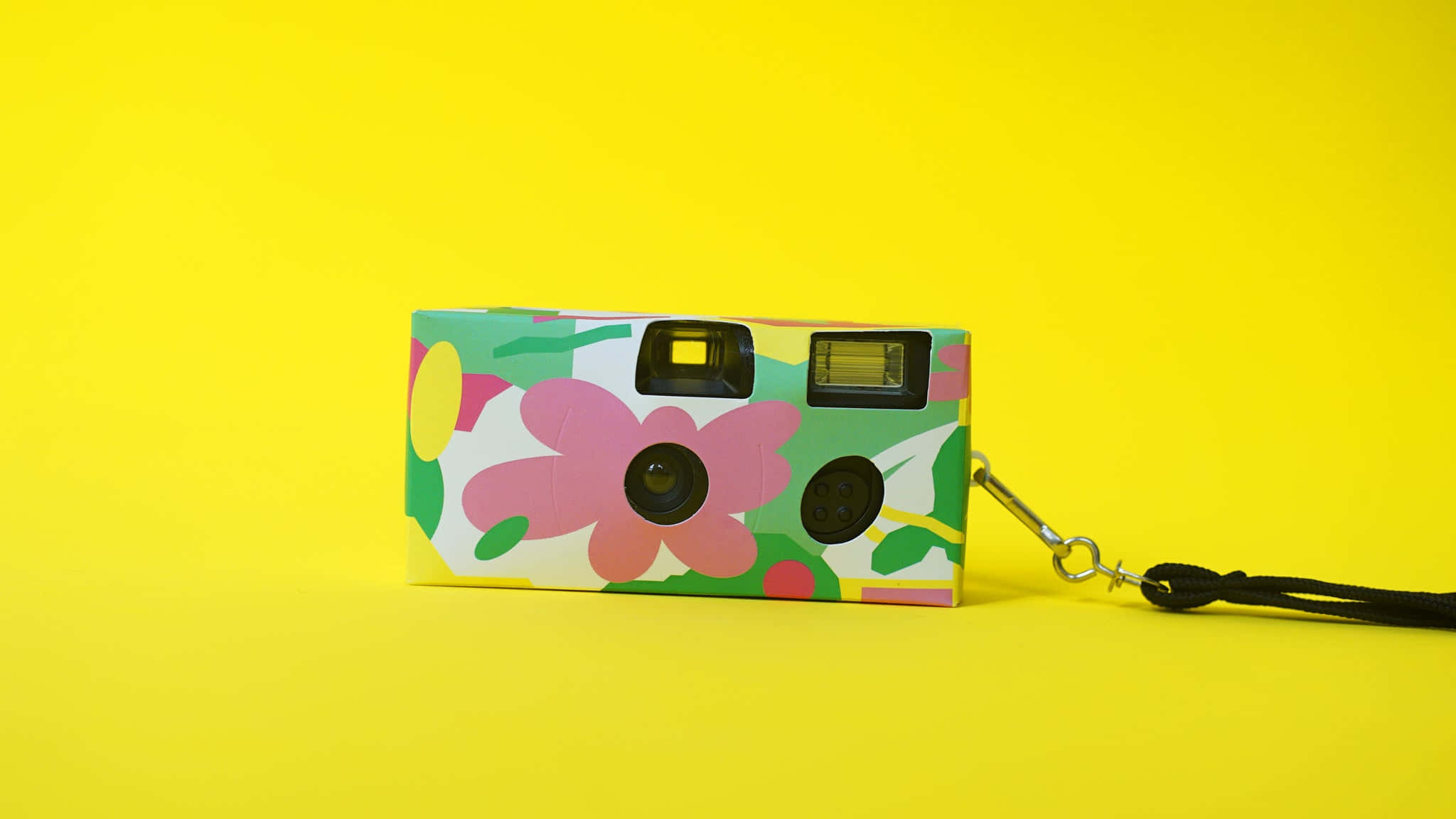 Disposable Camera 桌布 壁紙 — 一部色彩繽紛的花卉拍立得相機放在亮黃色背景上，配有黑色手腕帶。