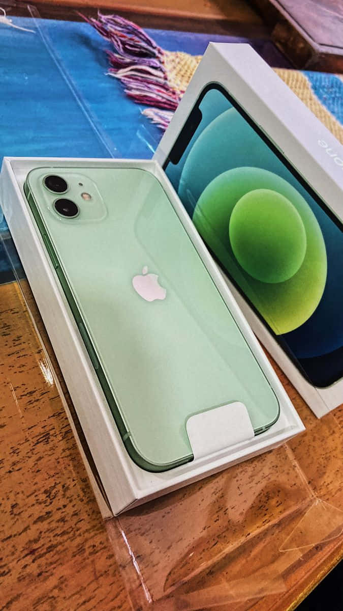 iPhone 12 桌布 壁紙 — 一部薄荷綠色的iPhone 12放在打開的盒子裡，置於木桌上，旁邊有色彩繽紛的編織布料。