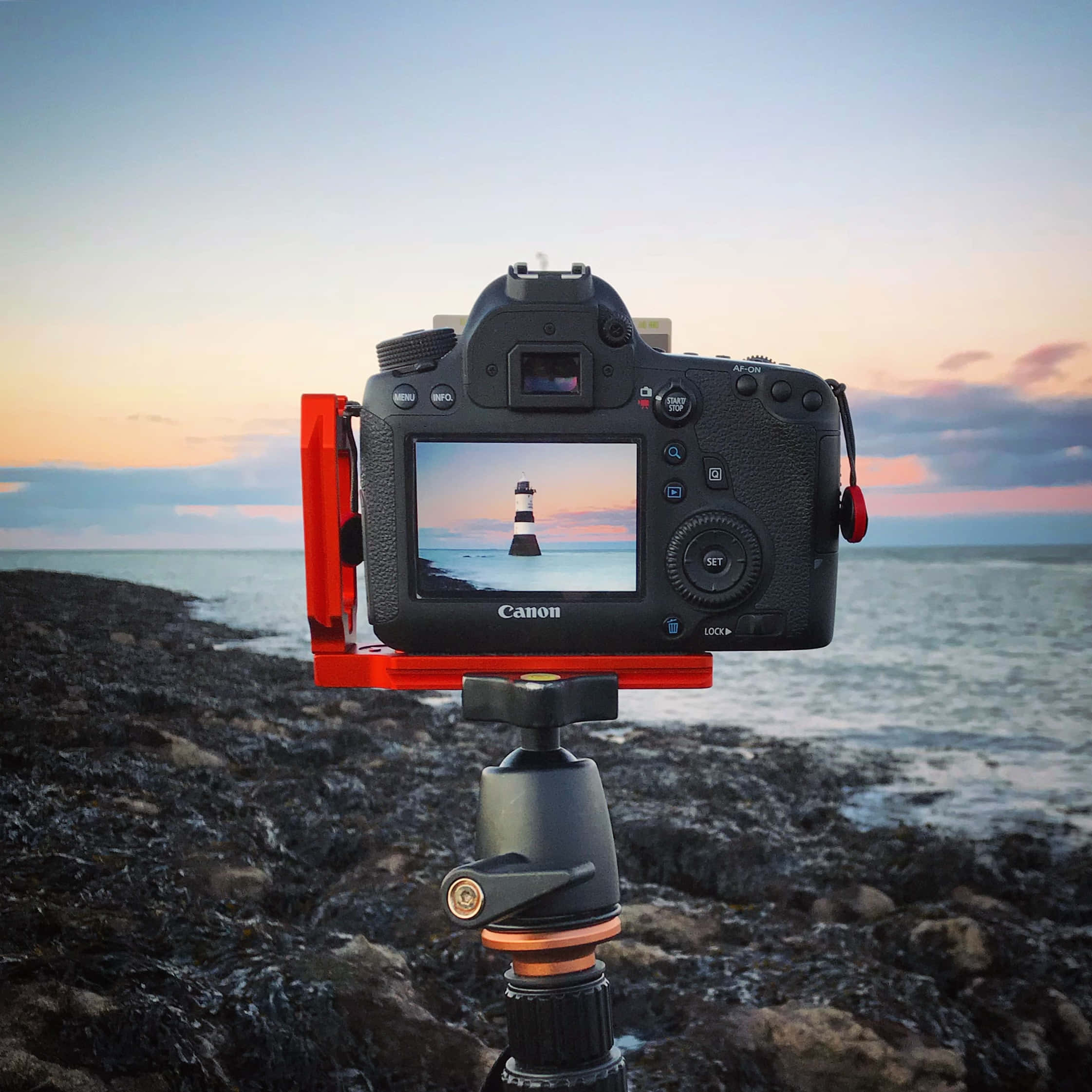 相機 桌布 壁紙 — 一部裝在三腳架上的Canon DSLR相機，捕捉日落時分岩石海岸線上的燈塔。