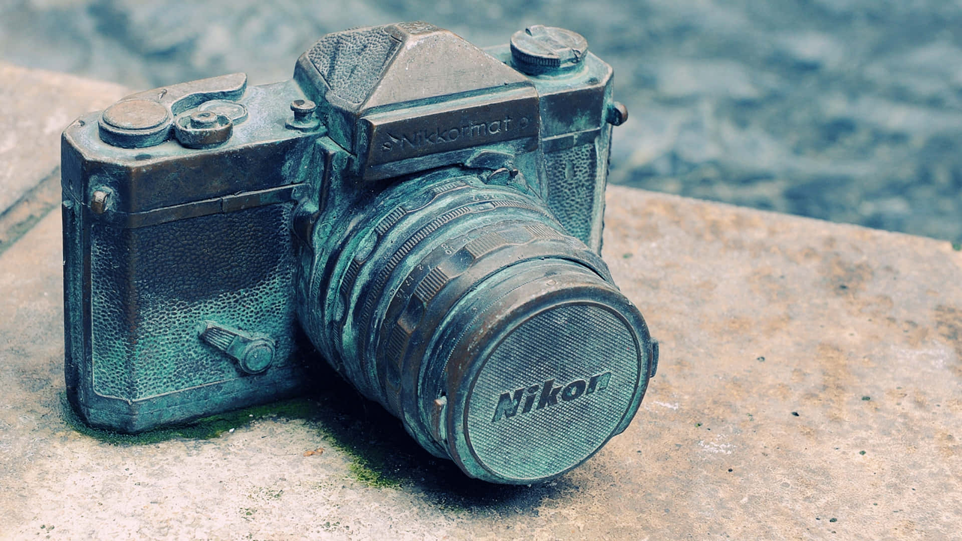 相機 桌布 壁紙 — 一部覆蓋著綠色銅綠的復古Nikon相機，擱在近水的風化石面上。