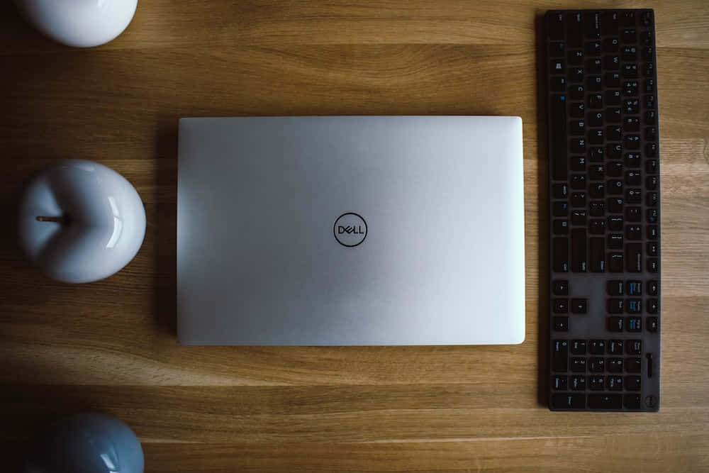 Dell 桌布 壁紙 — 一部銀色 Dell 筆記型電腦和黑色鍵盤放在溫暖的木桌上，旁邊有白色陶瓷蘋果形狀的物件。