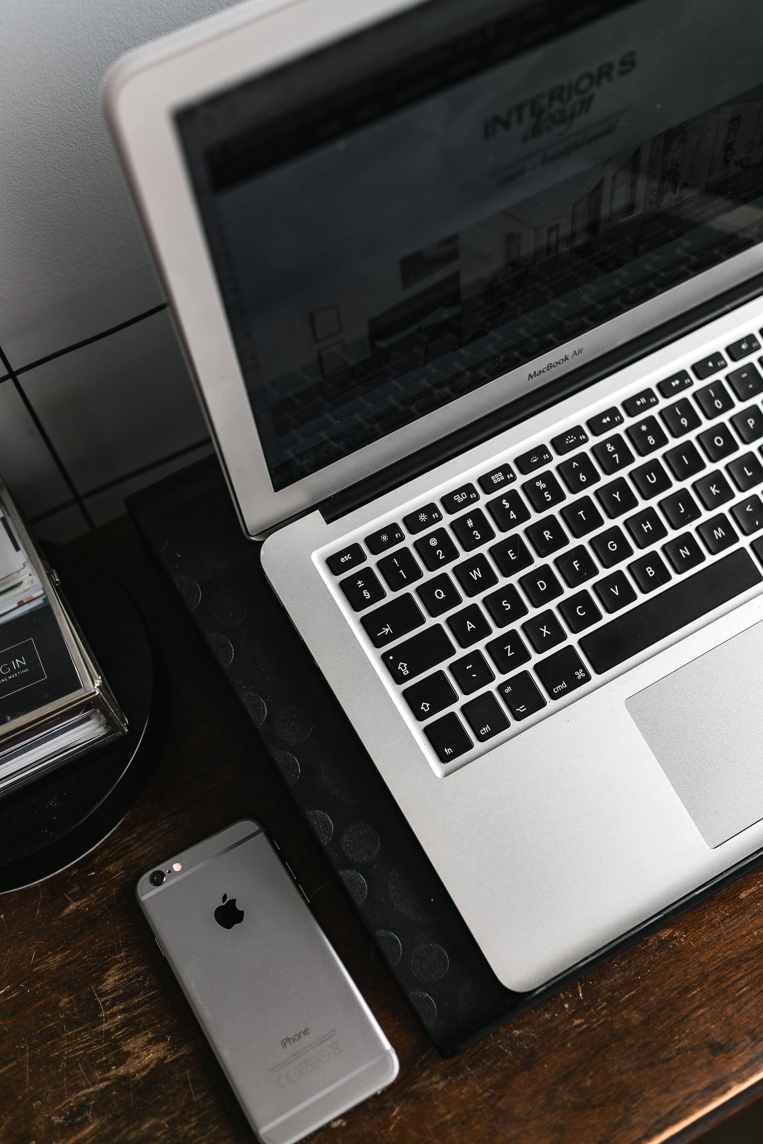 MacBook 動態圖片 桌布 壁紙 — 一部銀色 MacBook Air 和 iPhone 放在深色木桌上，旁邊是黑色磁磚牆和設計書籍。