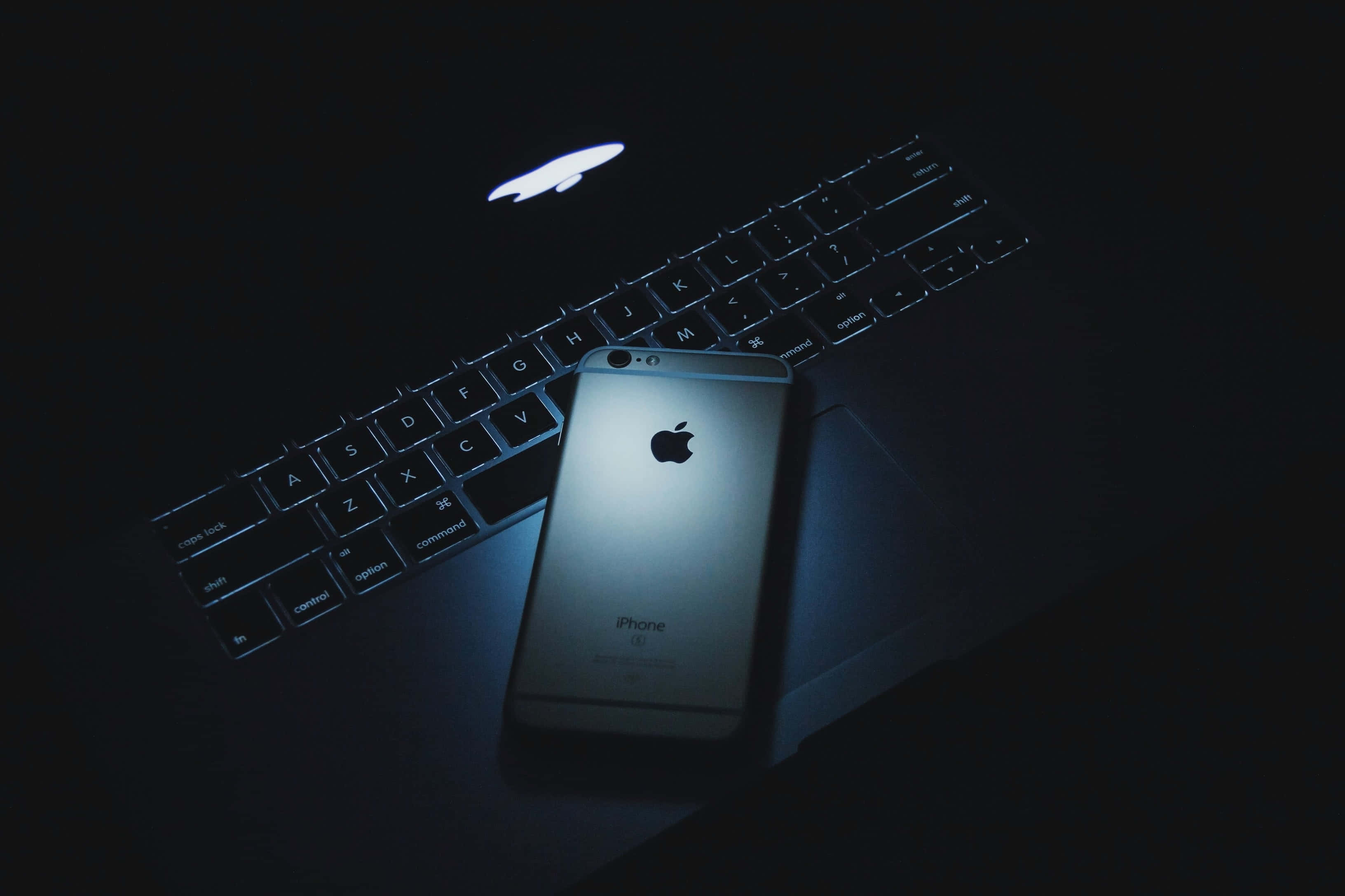 美學 MacBook 壁紙 桌布 壁紙 — 一部銀色iPhone在冷藍色燈光下放在深色筆記型電腦鍵盤上，Apple標誌發光。