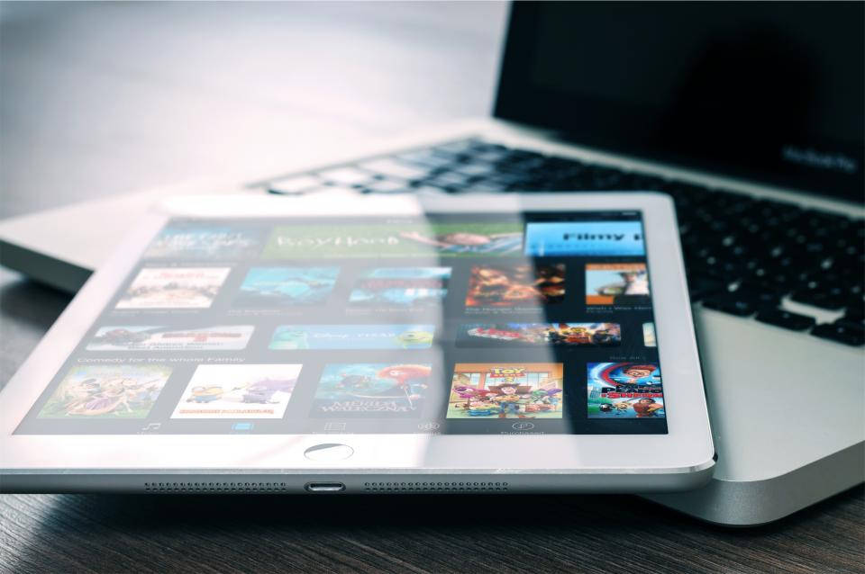 Apple iPad Pro 桌布 壁紙 — 一部銀色平板電腦顯示色彩繽紛的電影縮圖，放在木桌上，旁邊有筆記型電腦鍵盤。