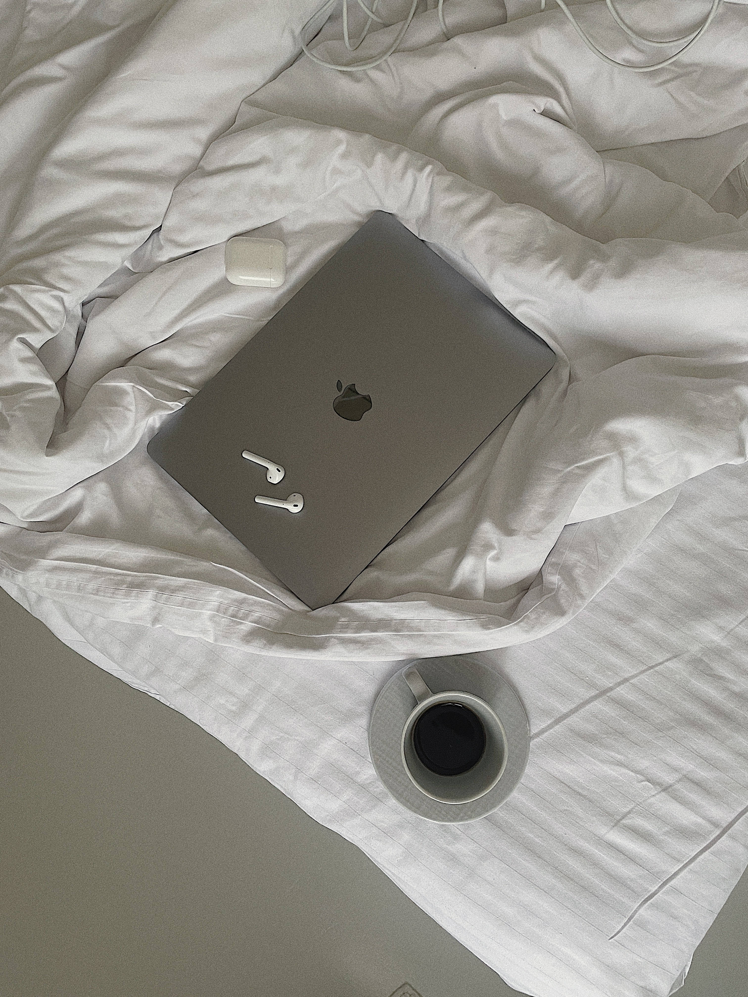 美學 桌布 壁紙 — 一部銀色筆記型電腦和白色耳機放在皺巴巴的白色床單上，旁邊有一杯黑咖啡。