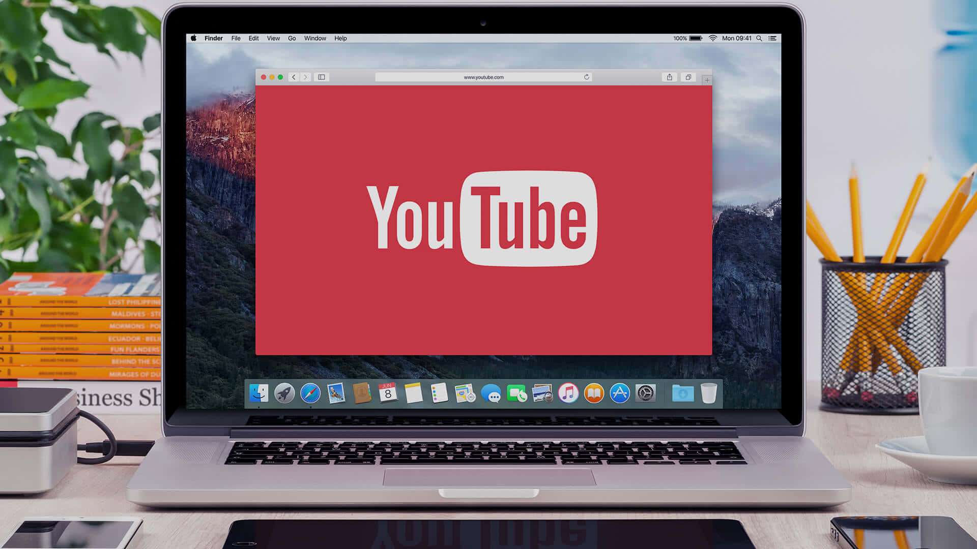 YouTube 桌布 壁紙 — 一部銀色筆記型電腦在紅色螢幕上顯示YouTube標誌，周圍有山脈桌面背景、辦公用品和植物。