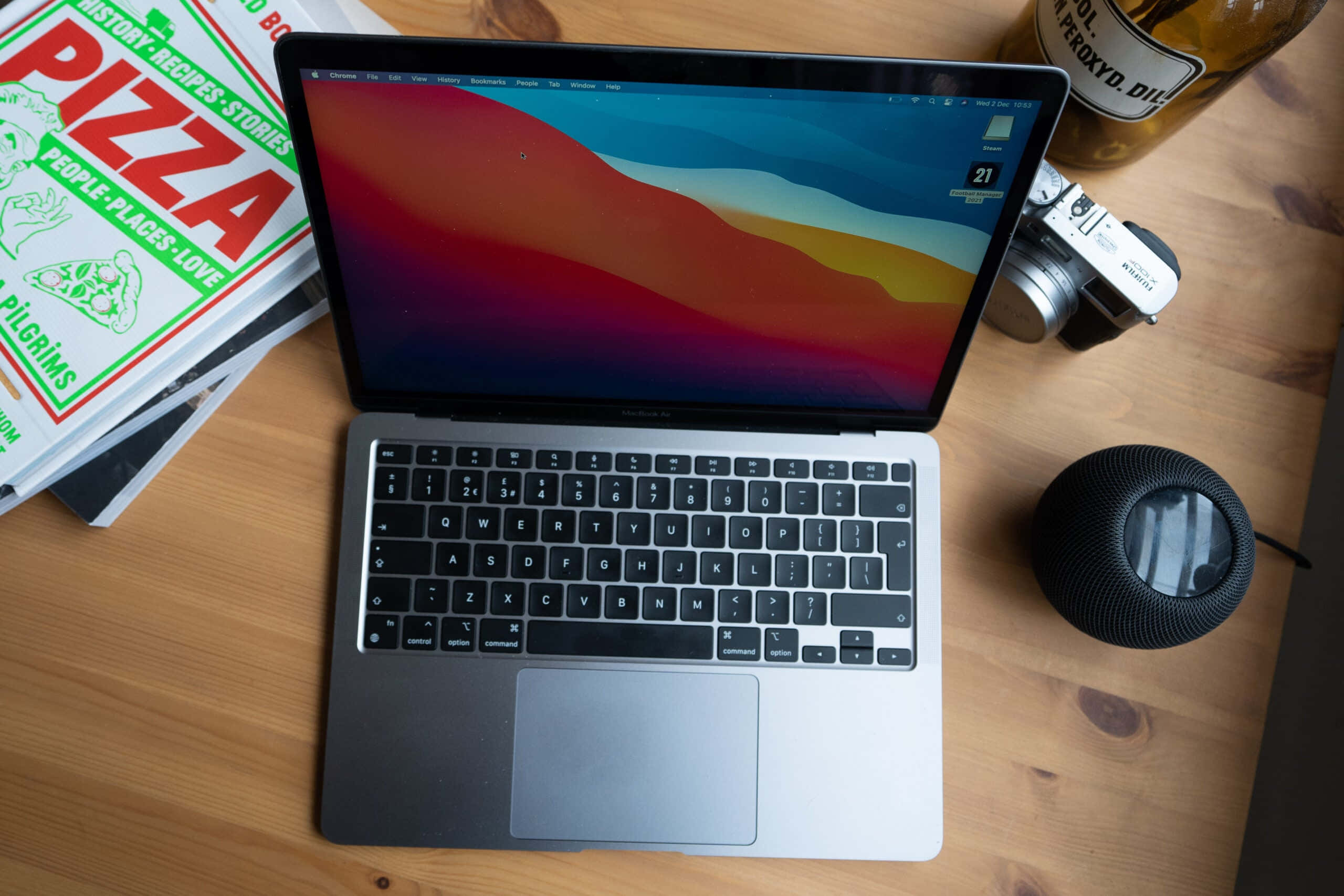 MacBook 動態圖片 桌布 壁紙 — 一部銀色筆記型電腦帶有彩色抽象桌布，放在木桌上，旁邊有相機、揚聲器和披薩書。