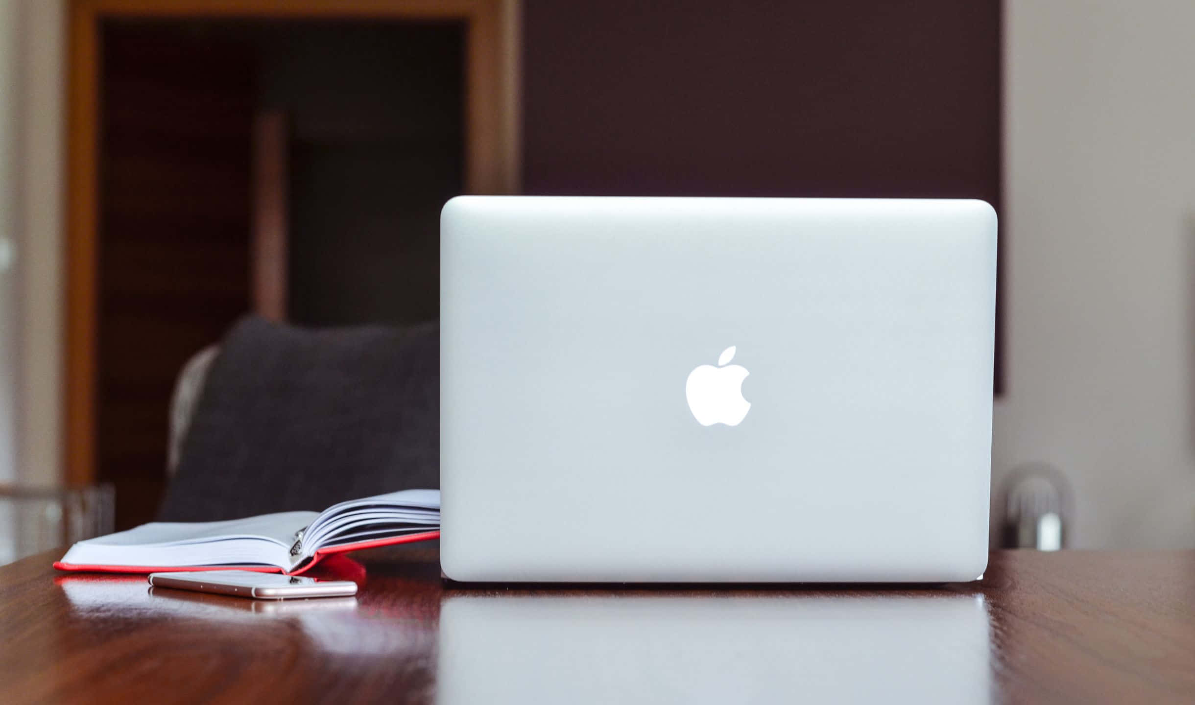2440x1440 Apple 桌布 壁紙 — 一部銀色筆記型電腦闔上放在木桌上，旁邊有一本打開的筆記本和智慧型手機，房間光線柔和。