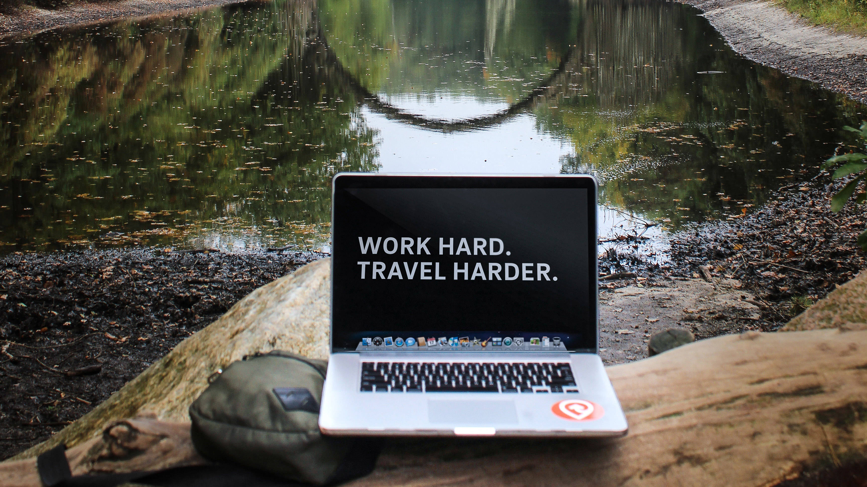 筆電 桌布 壁紙 — 一部銀色筆記型電腦顯示「WORK HARD. TRAVEL HARDER.」，擱在平靜森林池塘旁的一根圓木上，池塘倒映著樹木。