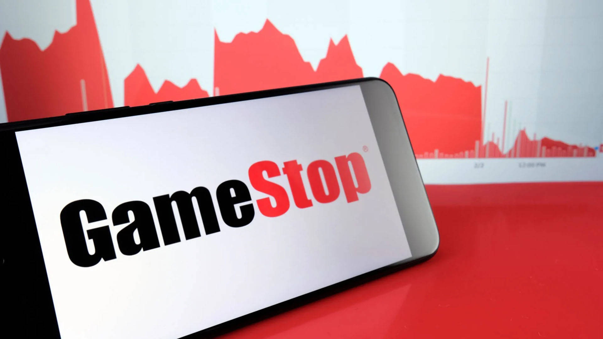GameStop 桌布 壁紙 — 一部顯示GameStop標誌的智慧型手機放在紅色表面上，背景有紅色的股票圖表。