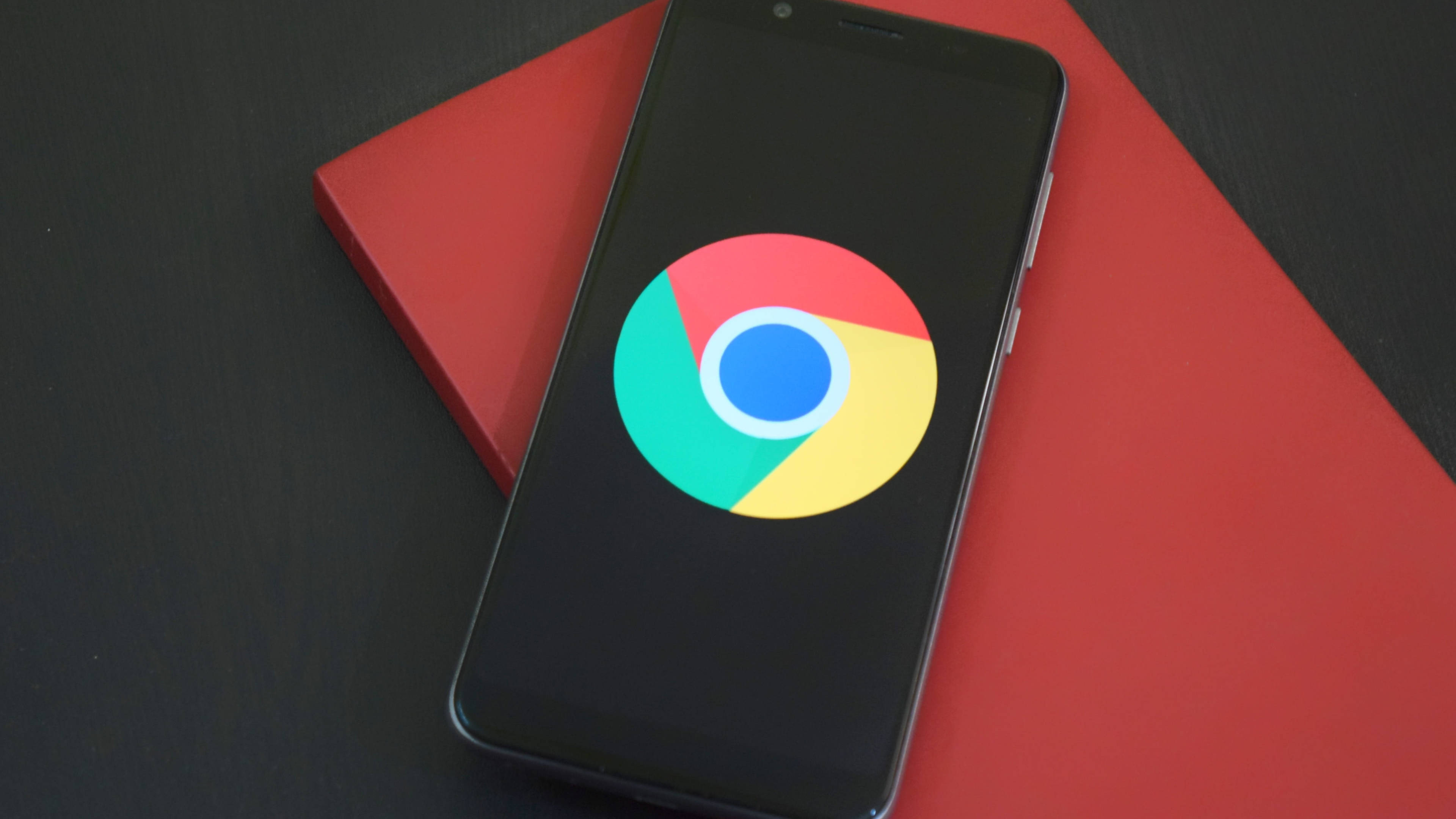 Google Chrome 桌布 壁紙 — 一部顯示Google Chrome標誌的智慧型手機放在深色表面上的鮮紅色書本上。