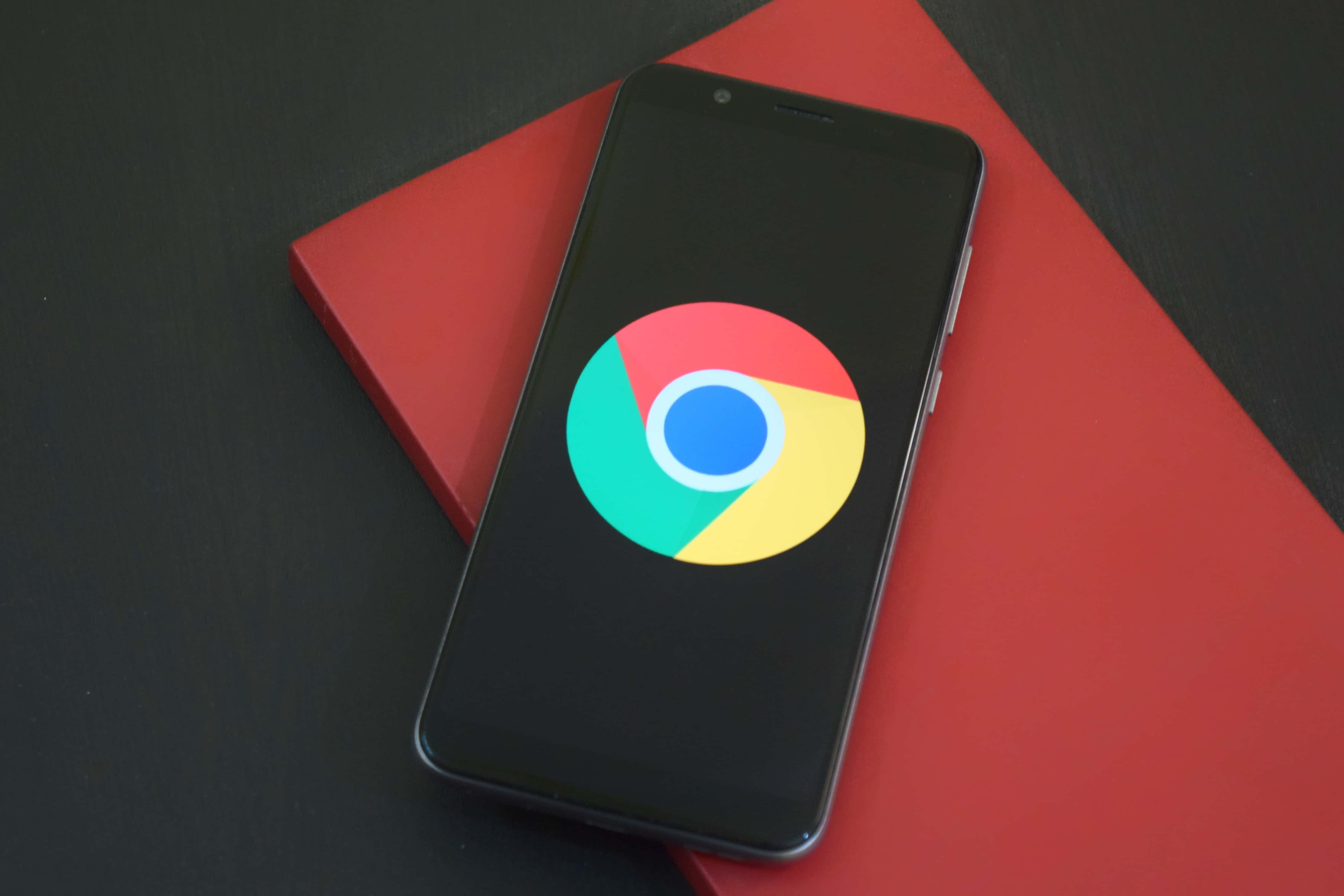 Chrome 桌布 壁紙 — 一部顯示Google Chrome標誌的智慧型手機放在深色表面上的鮮紅色筆記本上。