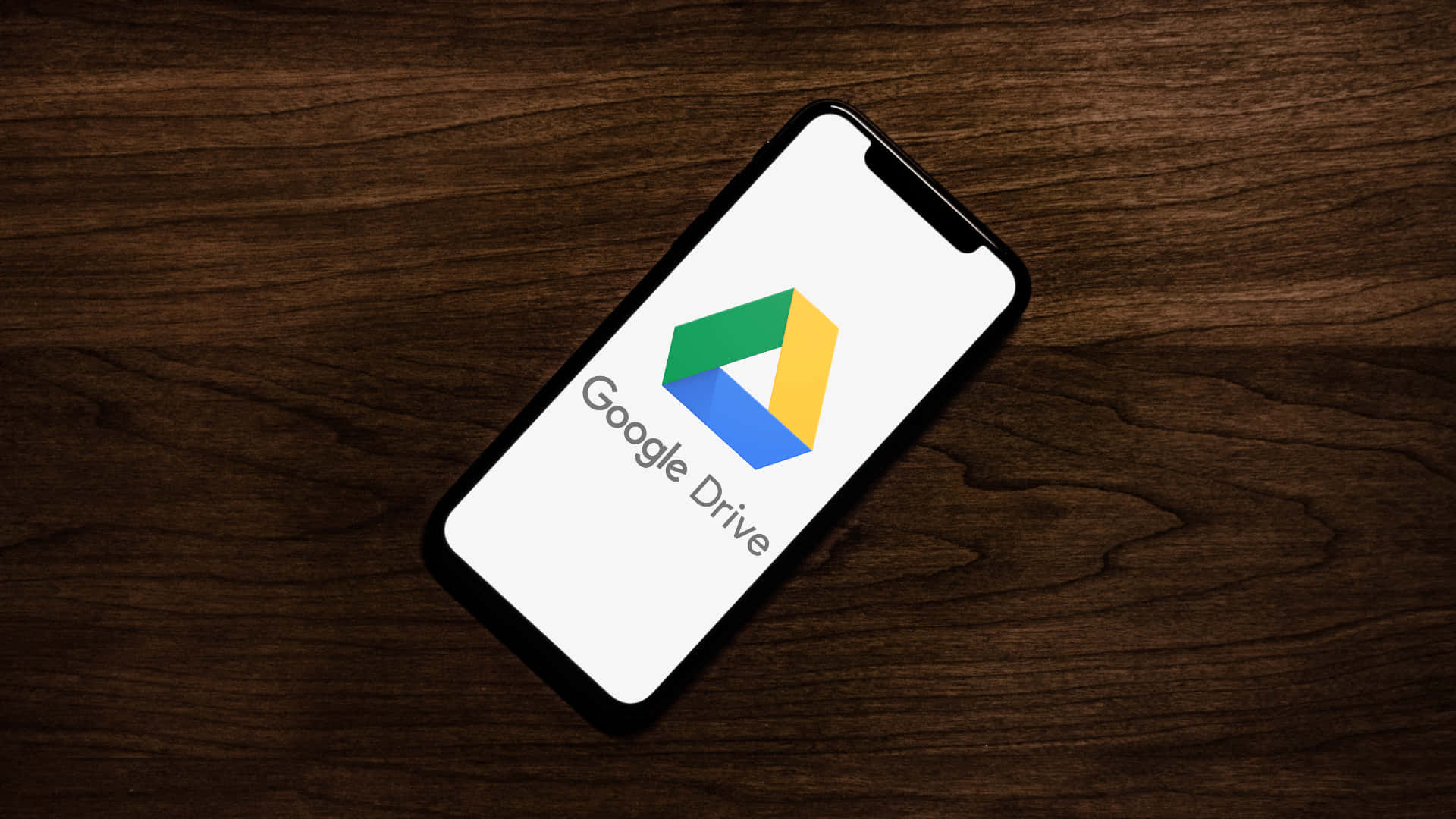 google 桌布 壁紙 — 一部顯示Google Drive標誌的智慧型手機對角放置在深色木質表面上。