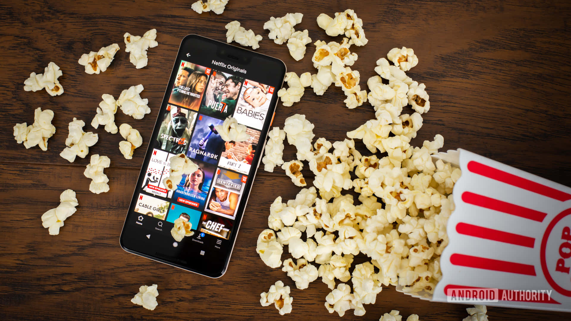 Netflix 桌布 壁紙 — 一部顯示Netflix Originals的智慧型手機，周圍散落著爆米花和一個紅白條紋桶，放在深色木桌上。