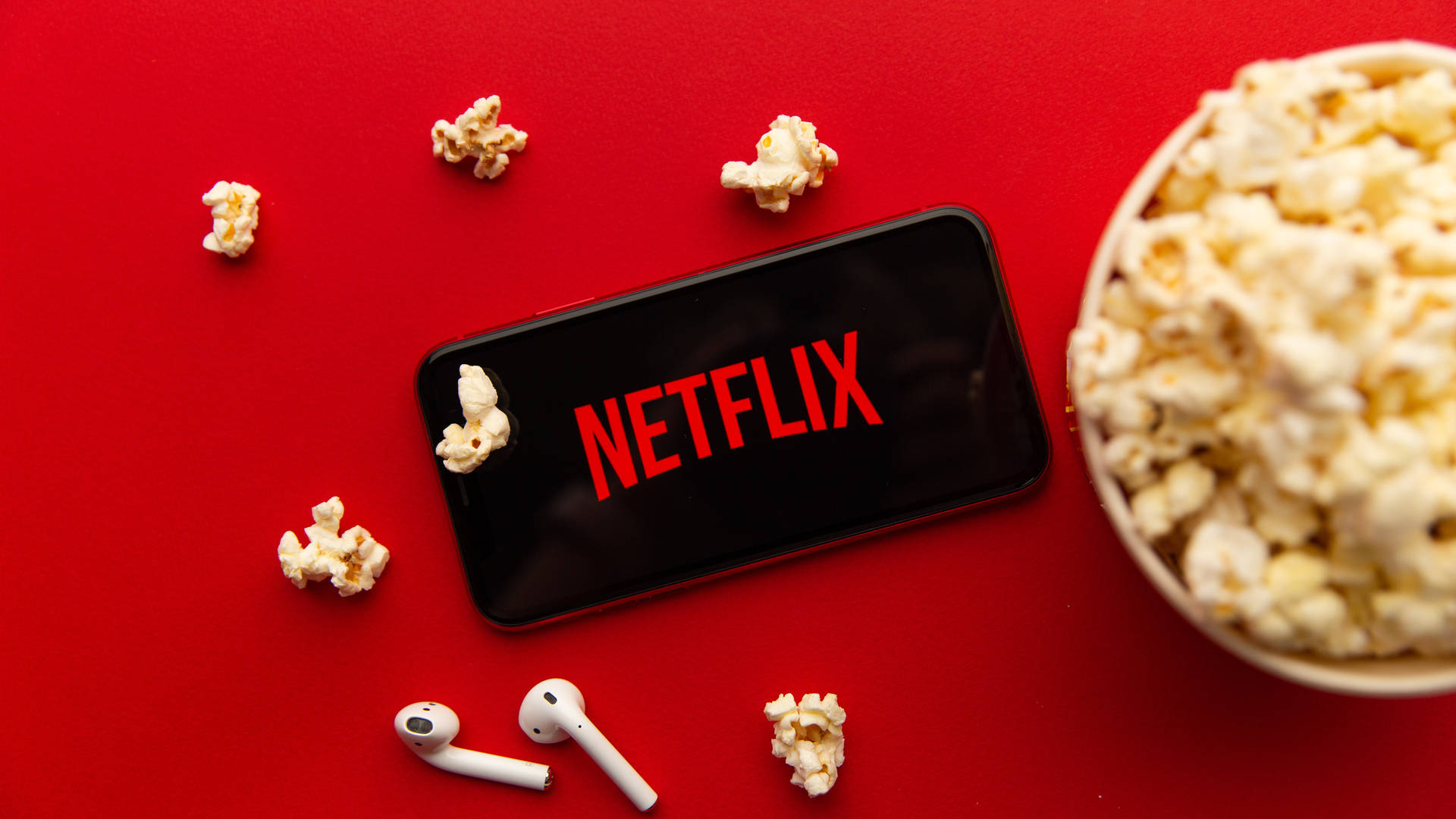 Netflix 桌布 壁紙 — 一部顯示Netflix標誌的智慧型手機放在鮮紅色的表面上，周圍散落著爆米花和白色無線耳機。
