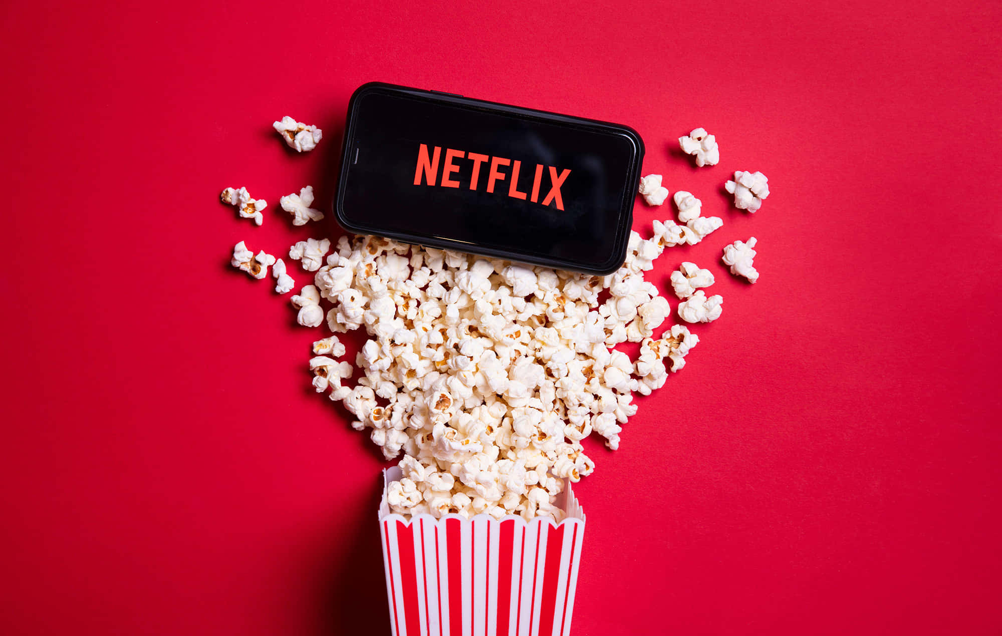 Netflix 桌布 壁紙 — 一部顯示Netflix標誌的智慧型手機放在一個滿溢的紅白條紋爆米花桶上，背景為鮮紅色。