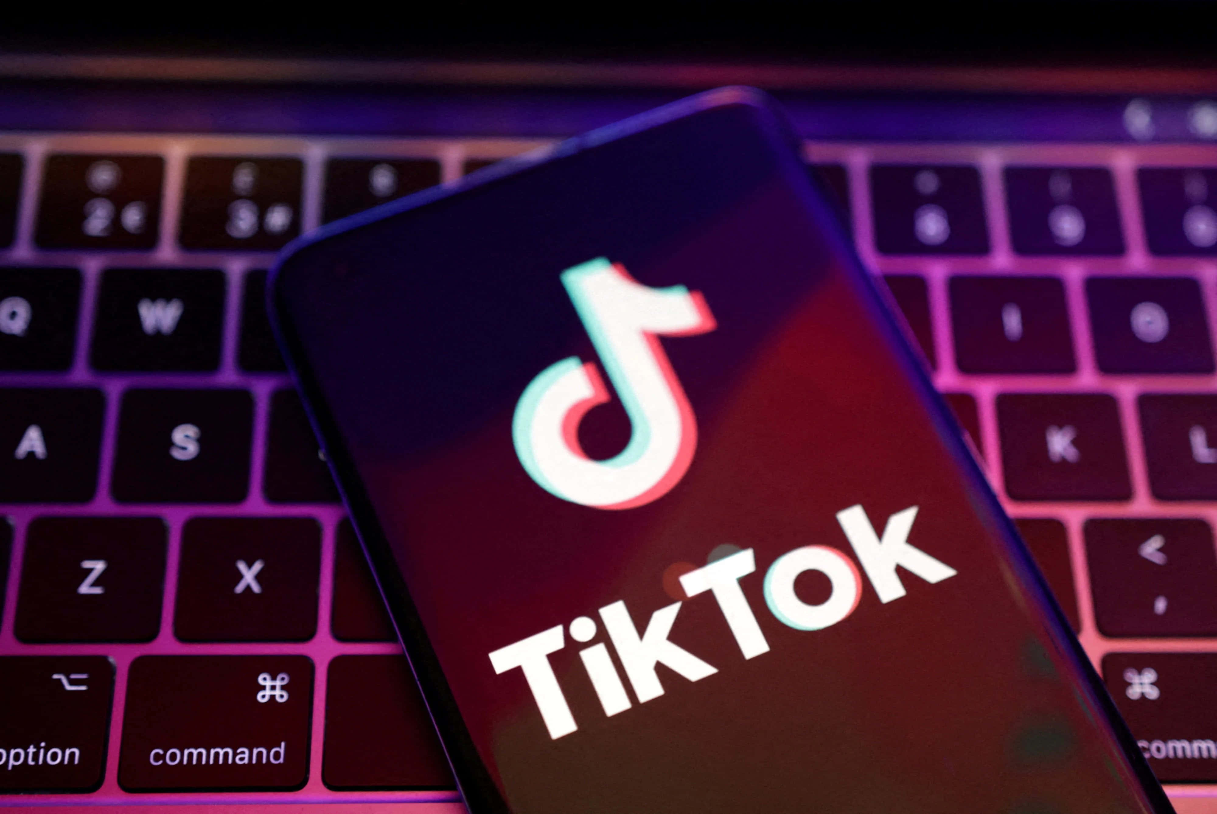 TikTok 動圖 桌布 壁紙 — 一部顯示TikTok標誌的智慧型手機放在一個沐浴在紫色和粉紅色霓虹燈光下的鍵盤上。