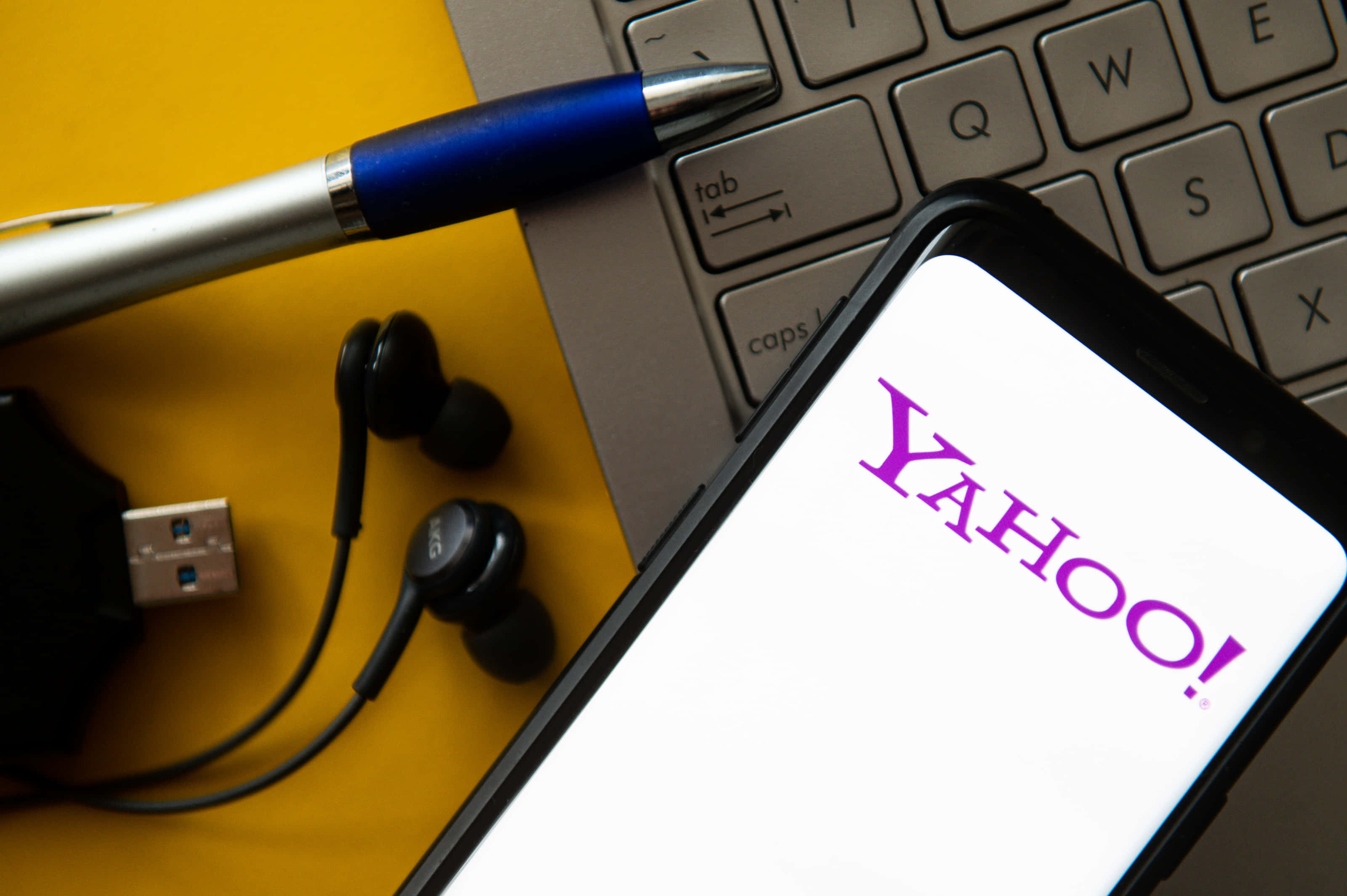 Yahoo 桌布 壁紙 — 一部顯示Yahoo標誌的智慧型手機放在一個鮮黃色表面上，旁邊有筆記型電腦鍵盤、藍色筆、耳塞和USB隨身碟。