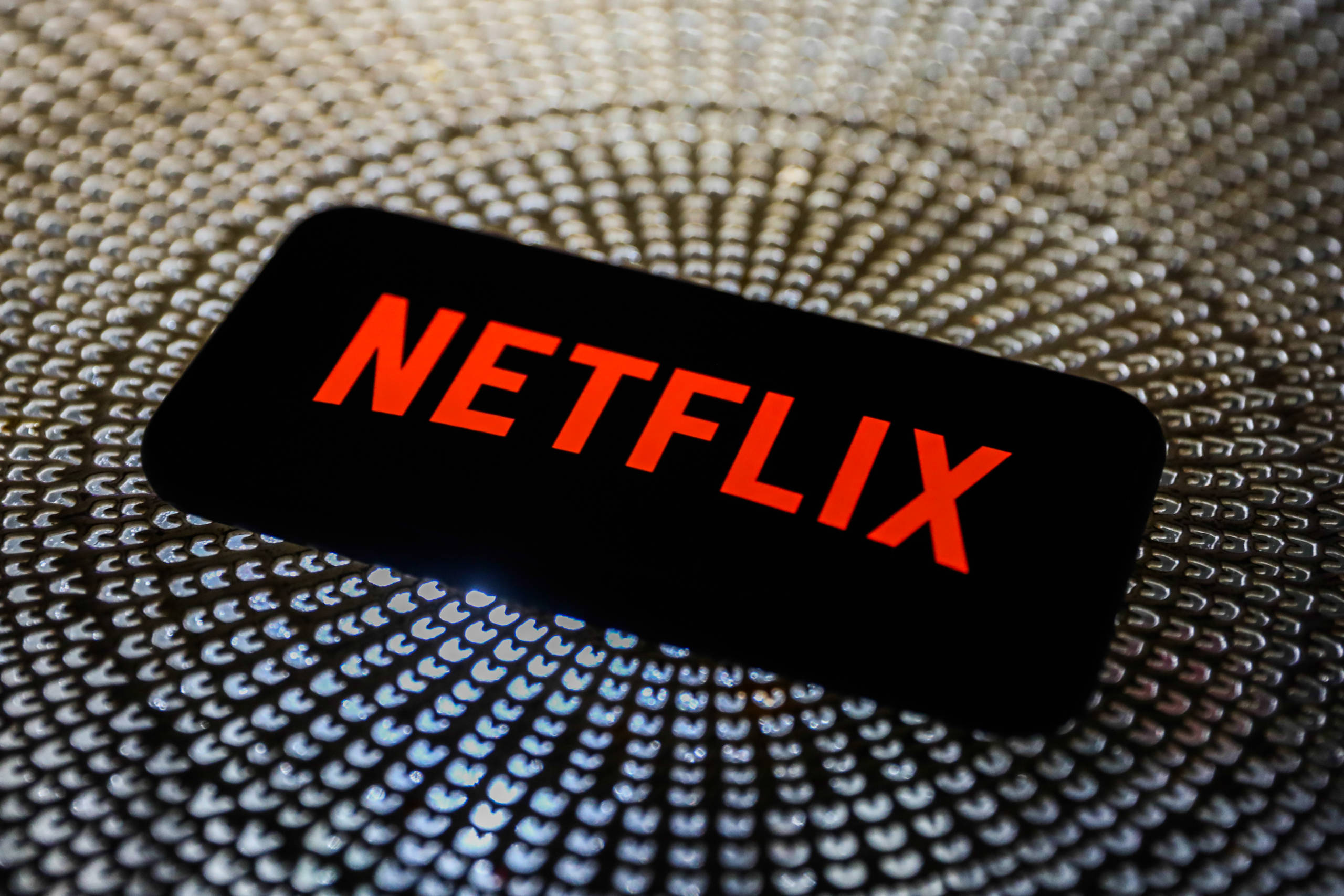 Netflix 桌布 壁紙 — 一部顯示亮紅色Netflix標誌的智慧型手機，放置在帶有放射狀圖案的紋理金屬表面上。