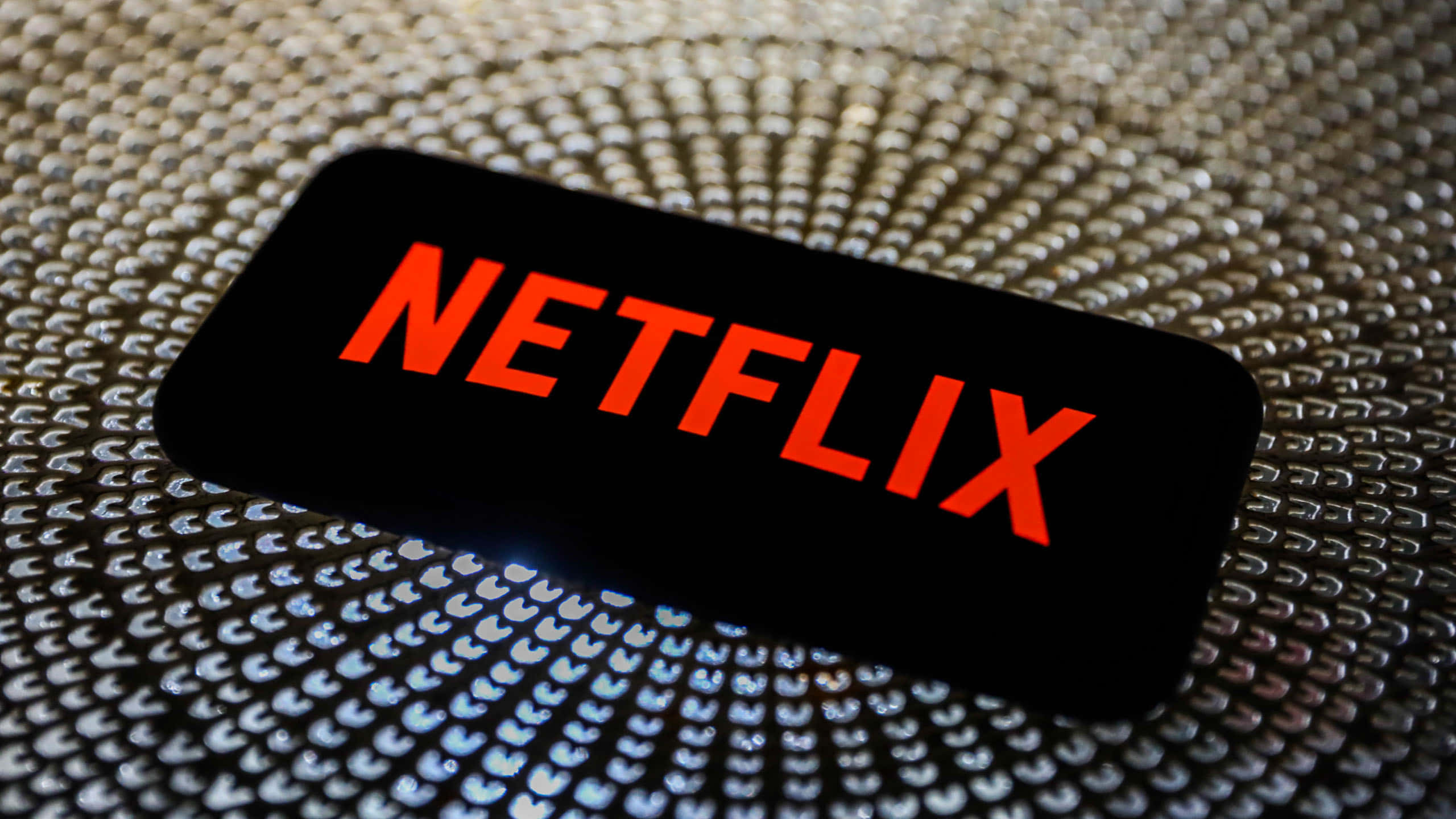 手機 桌布 壁紙 — 一部顯示亮紅色Netflix標誌的智慧型手機，放置在帶有放射狀圖案的紋理金屬表面上。