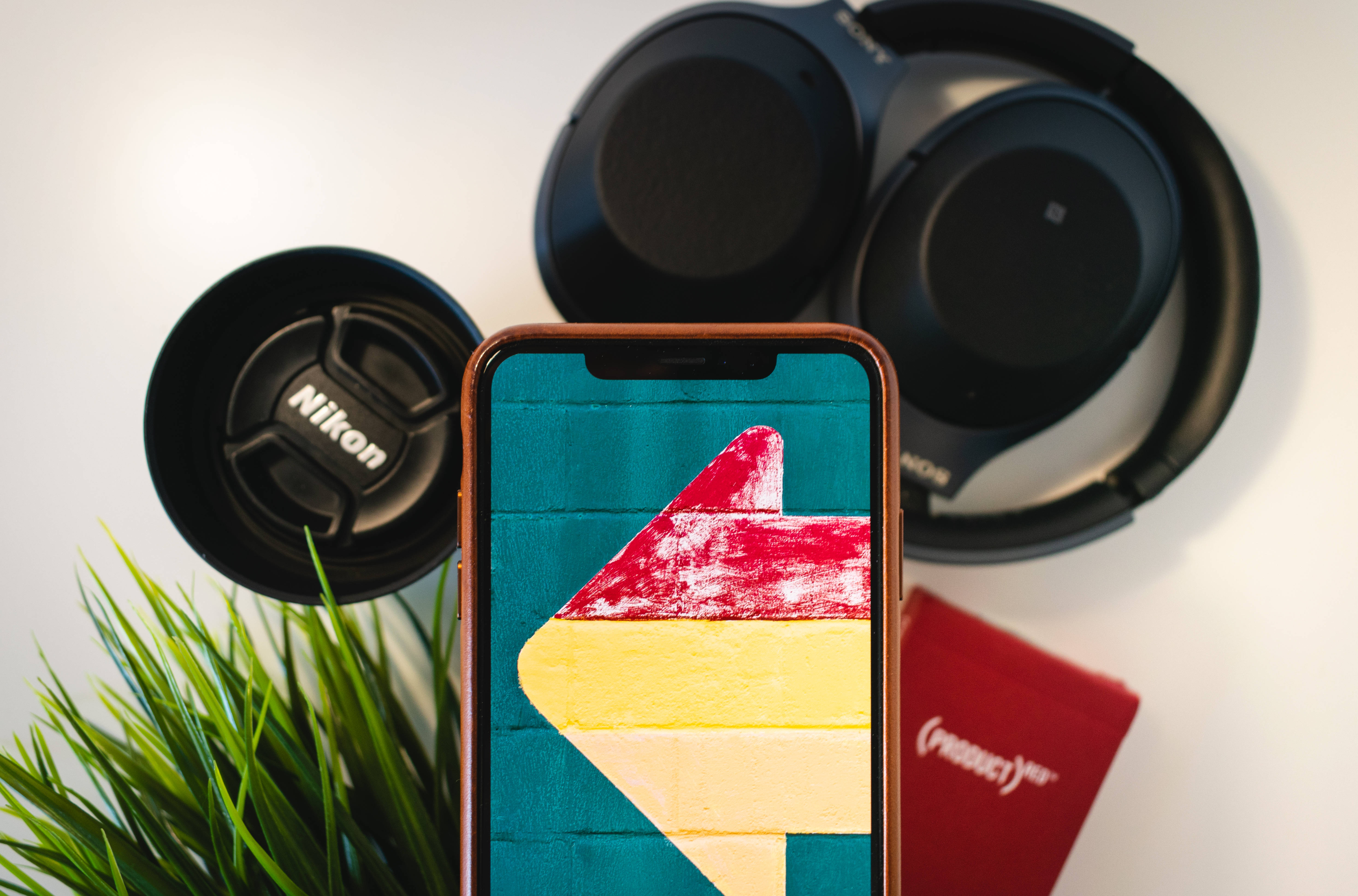 iPhone Xs Max 桌布 壁紙 — 一部顯示彩色抽象箭頭桌布的智慧型手機，與耳機、Nikon鏡頭蓋和綠色植物一起放在白色表面上。