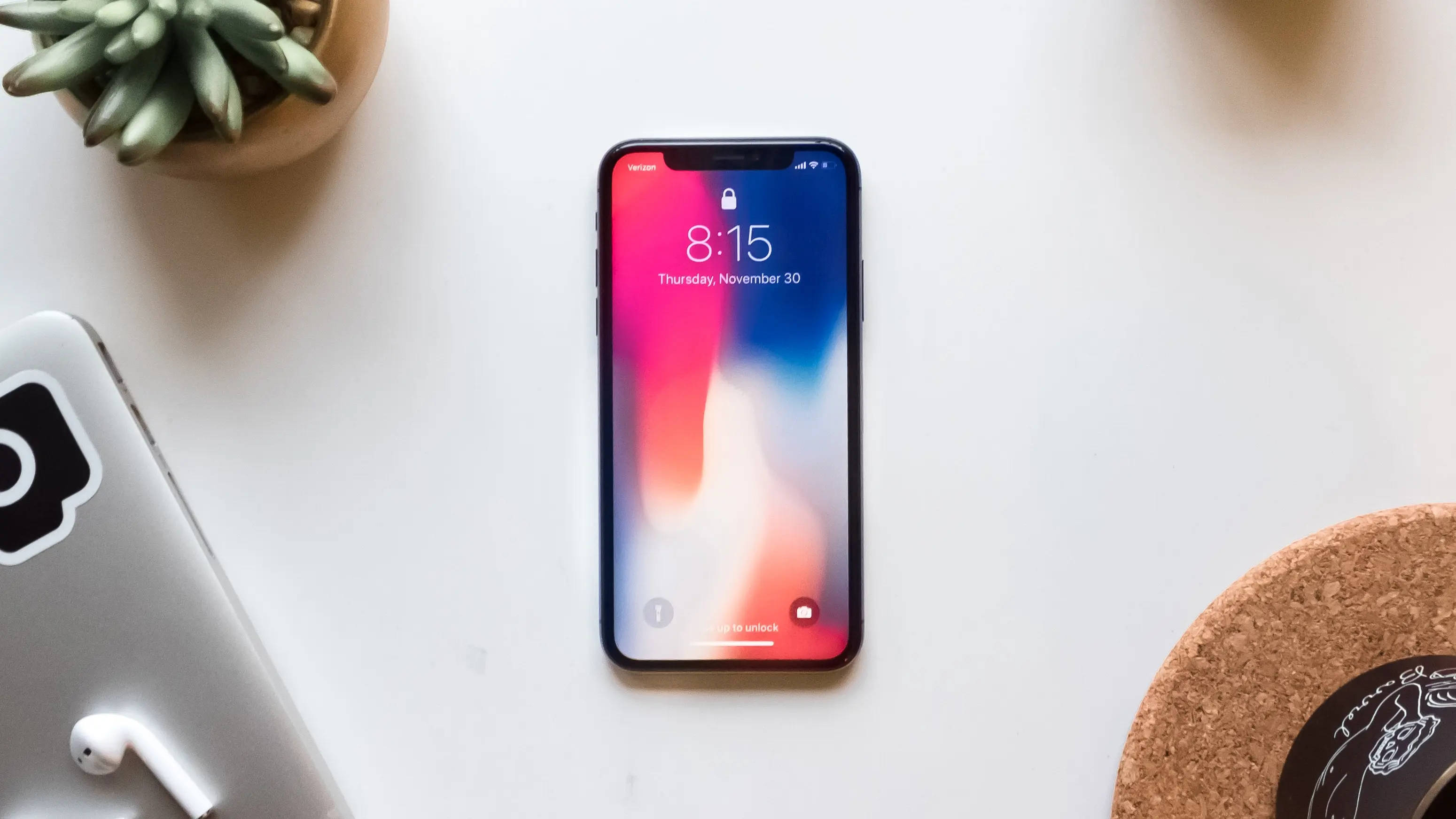 Interesting iPhone 桌布 壁紙 — 一部顯示彩色漸層桌布的智慧型手機放在白色書桌中央，旁邊有一盆多肉植物和耳塞。