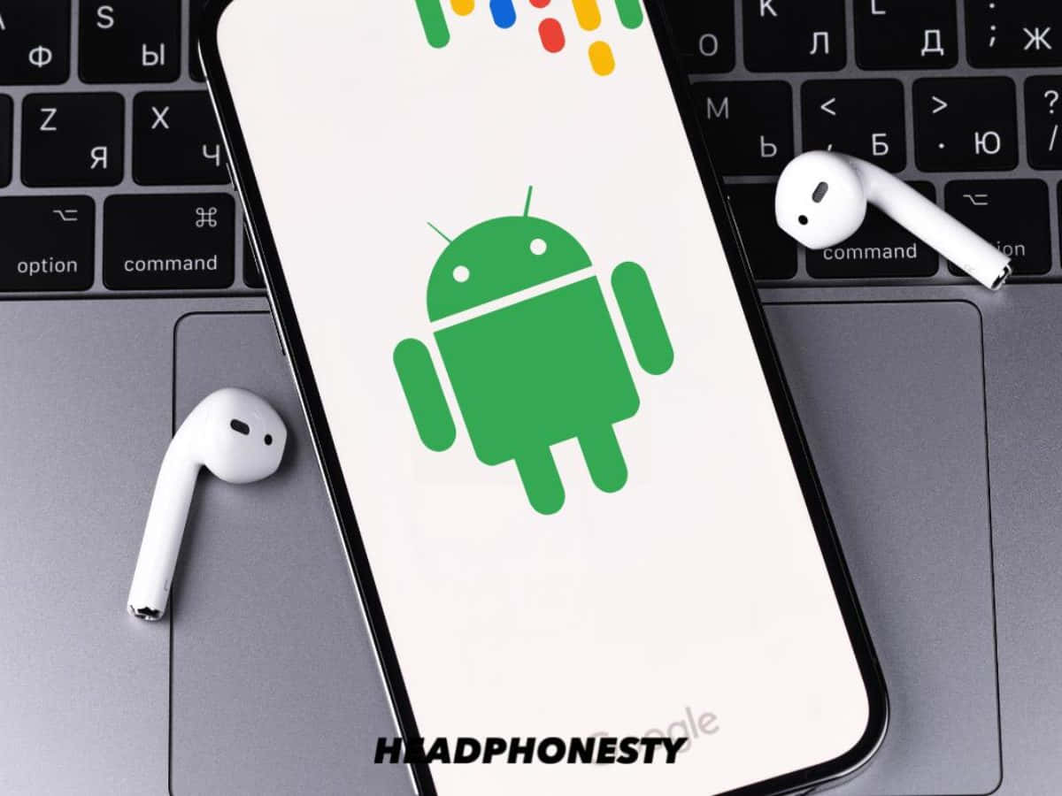 安卓 桌布 壁紙 — 一部顯示綠色Android吉祥物的智慧型手機放在筆記型電腦鍵盤上，旁邊有白色無線耳機。