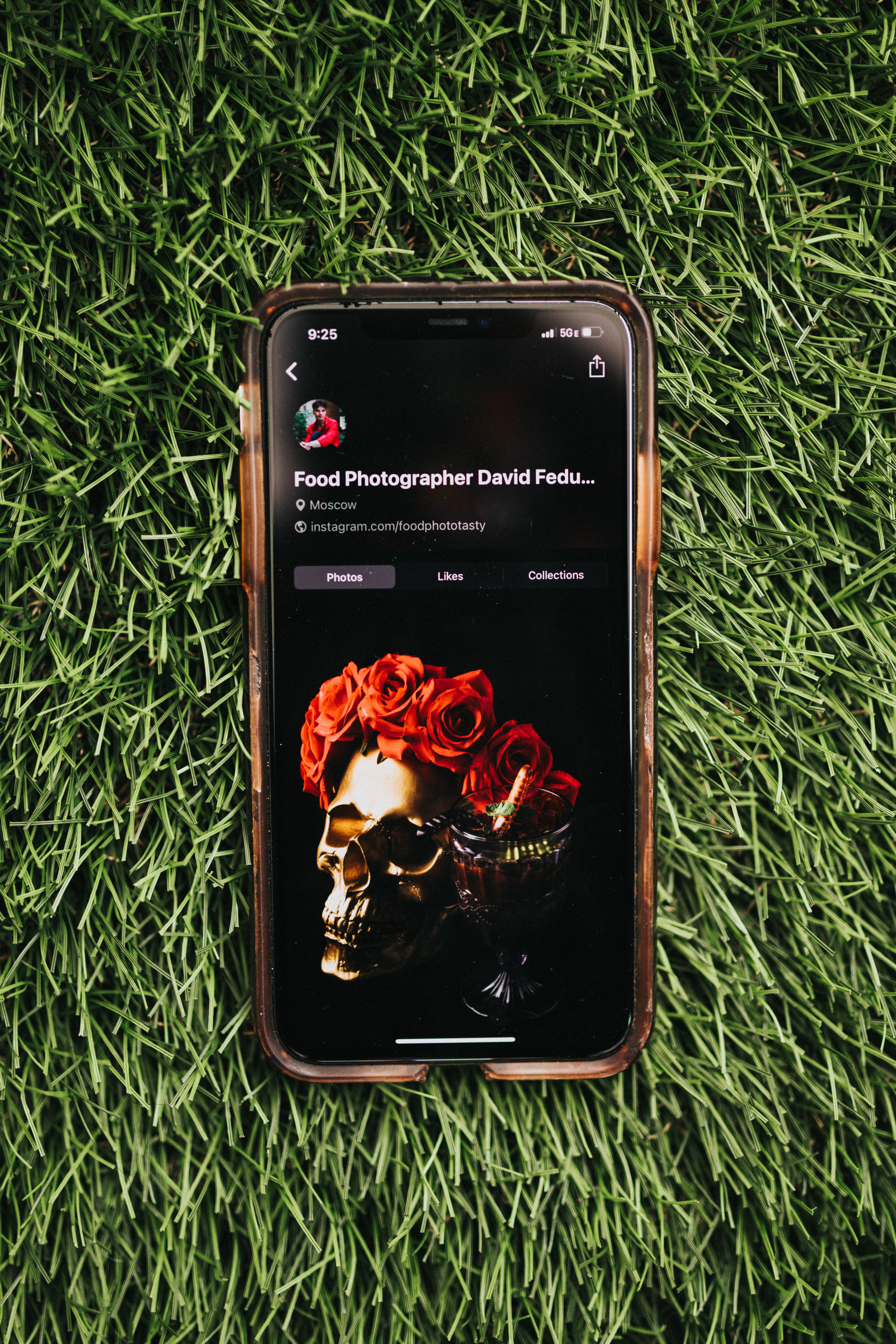 iOS 桌布 壁紙 — 一部顯示美食攝影師Instagram個人資料的智慧型手機放在鮮綠色草地上，展示一個飾有紅玫瑰的金色頭骨。