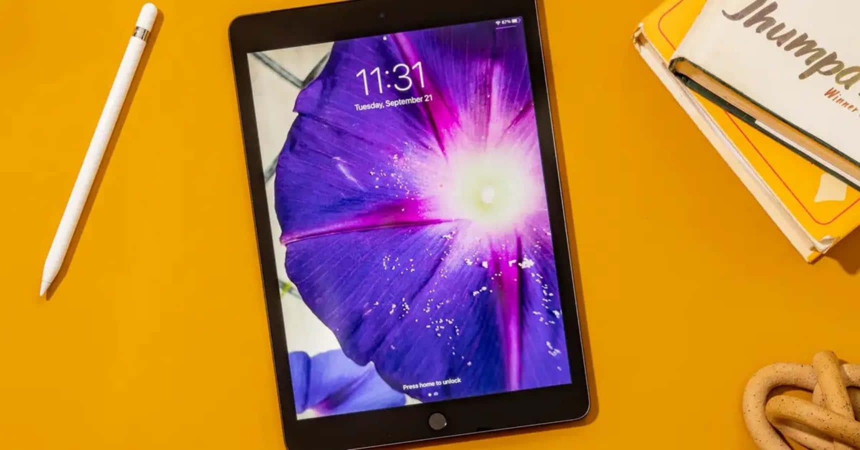 iPad 桌布 壁紙 — 一部顯示鮮豔紫色花朵桌面的平板電腦，放置在亮黃色表面上，旁邊有觸控筆和書籍。