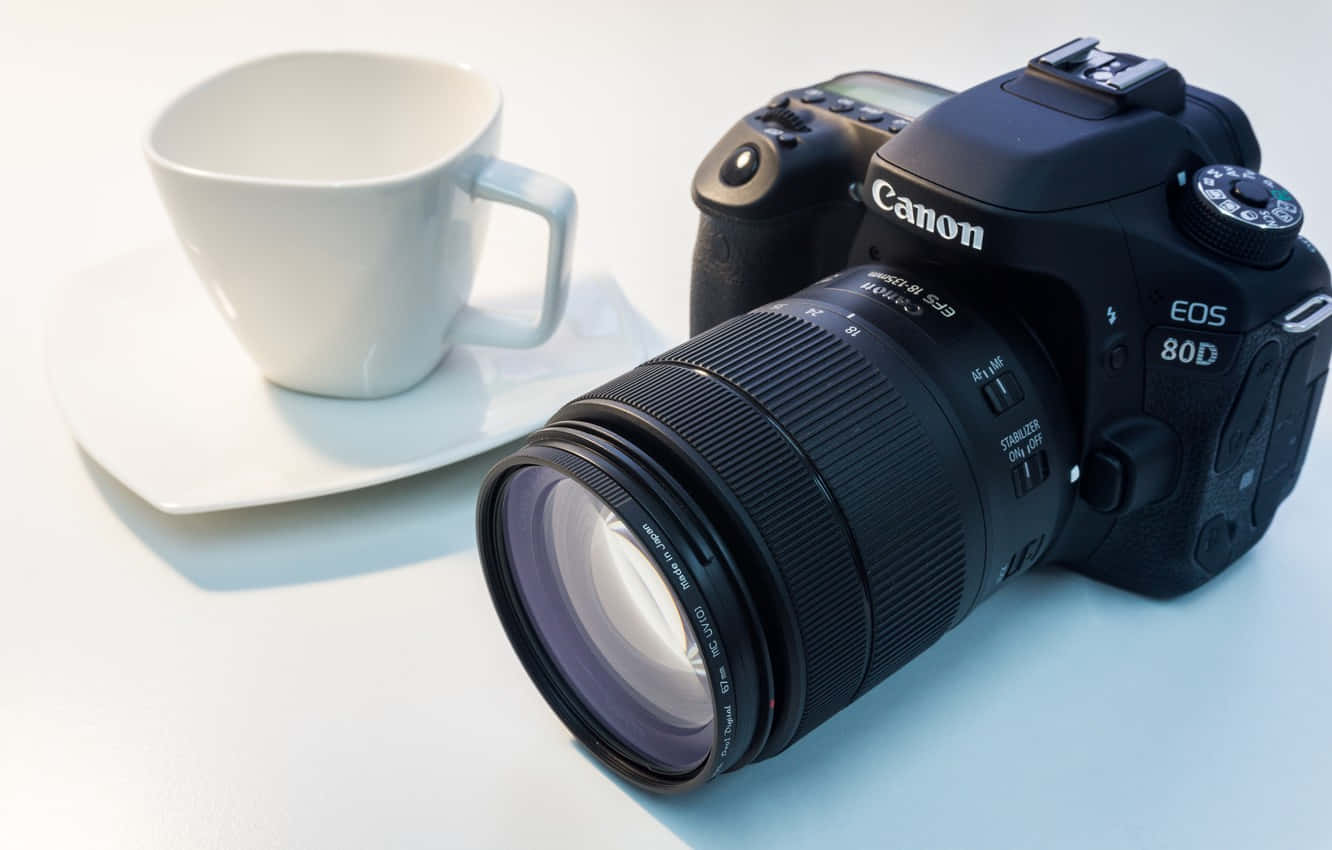 Dslr 桌布 壁紙 — 一部黑色 Canon EOS 80D DSLR 相機連鏡頭放在白色陶瓷杯和碟旁邊，背景是純白色。