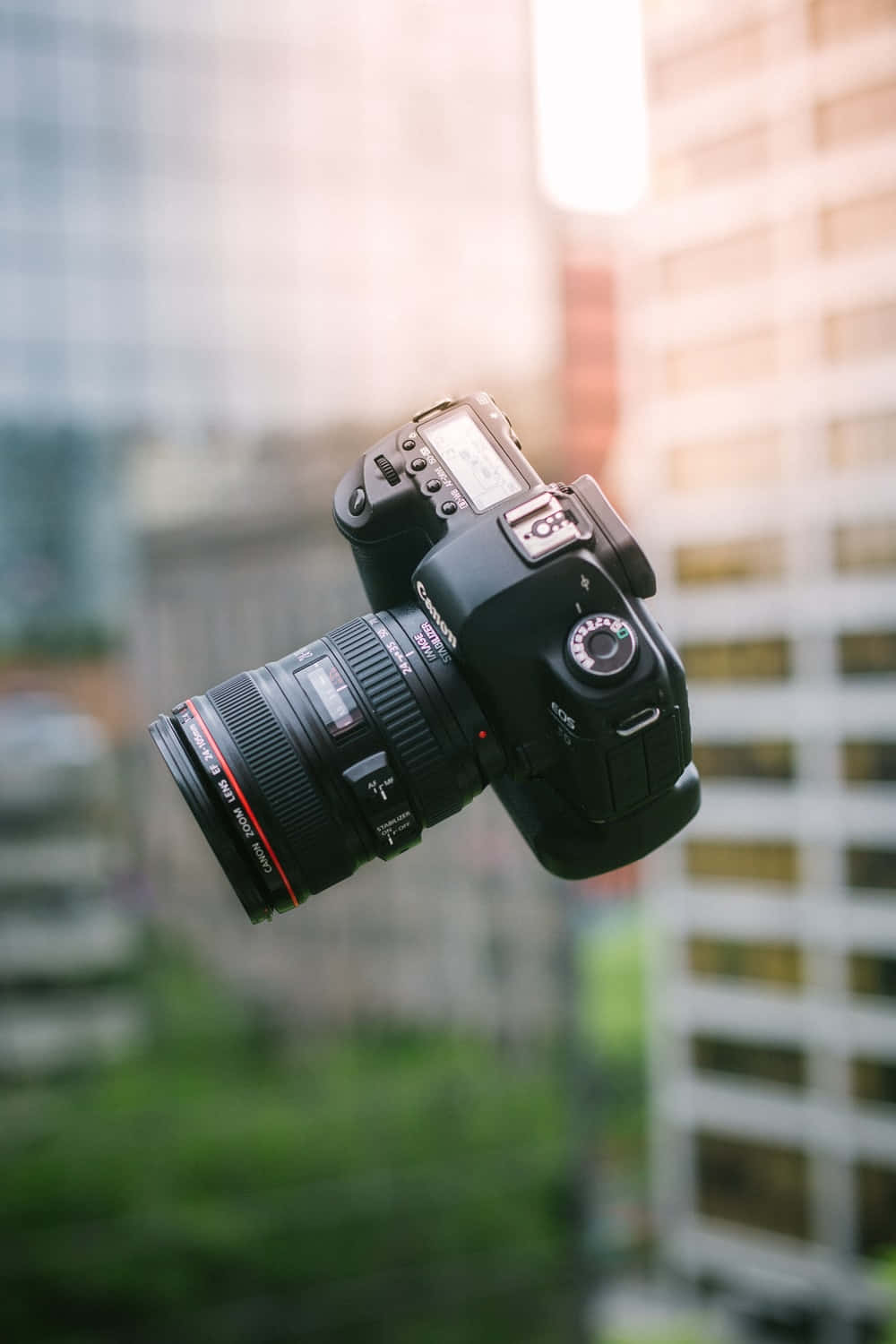 Dslr 桌布 壁紙 — 一部黑色 DSLR 相機在半空中漂浮，背景是模糊的城市景觀，溫暖的金色光線穿過建築物。