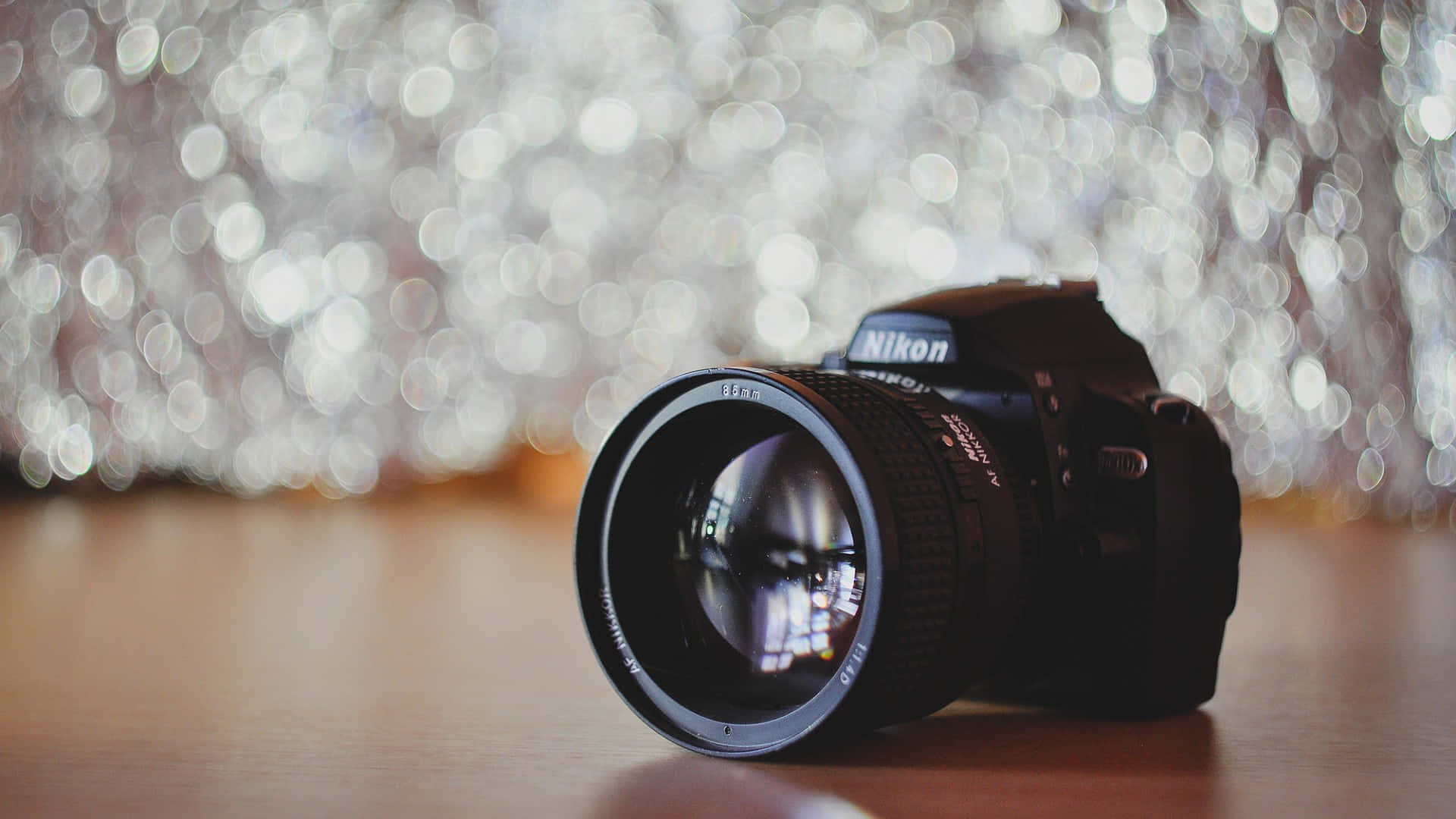 相機 桌布 壁紙 — 一部黑色 Nikon DSLR 相機放在木質表面上，背景有閃爍的散景燈光。
