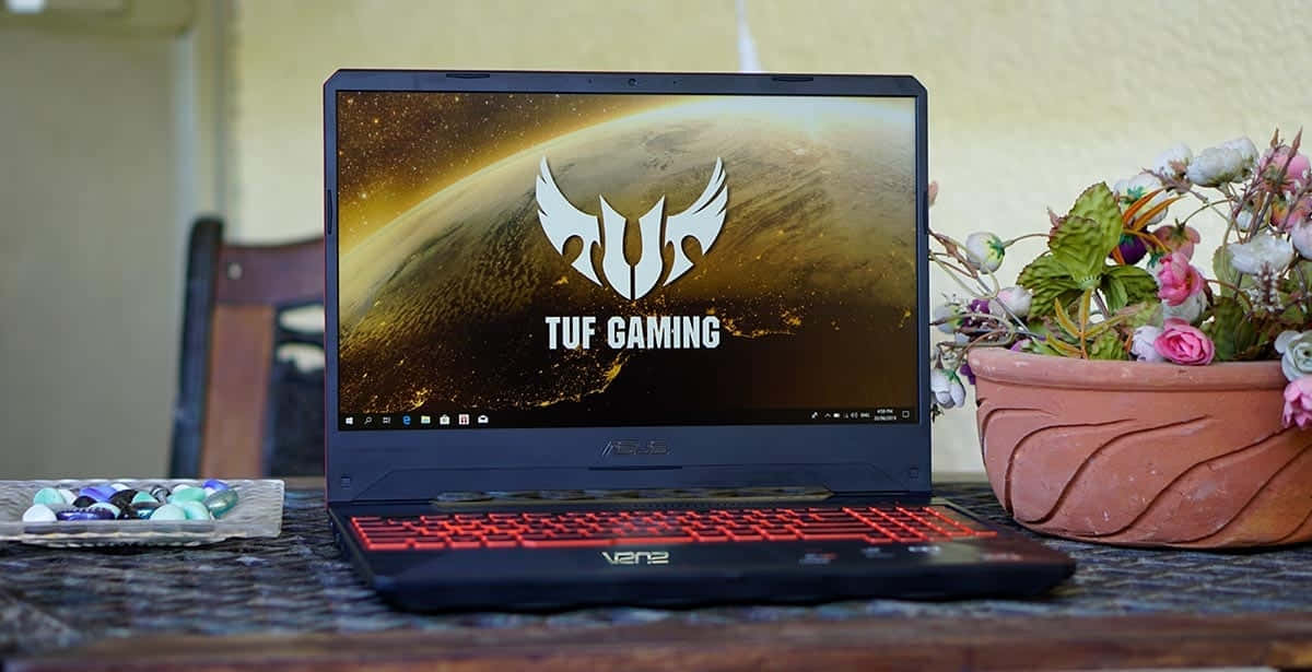 ASUS 桌布 壁紙 — 一部黑色ASUS TUF Gaming筆記型電腦，帶有紅色背光按鍵，打開放在木桌上，旁邊有一個赤陶花盆，裡面有粉紅色和白色的花，還有一盤色彩繽紛的彈珠。